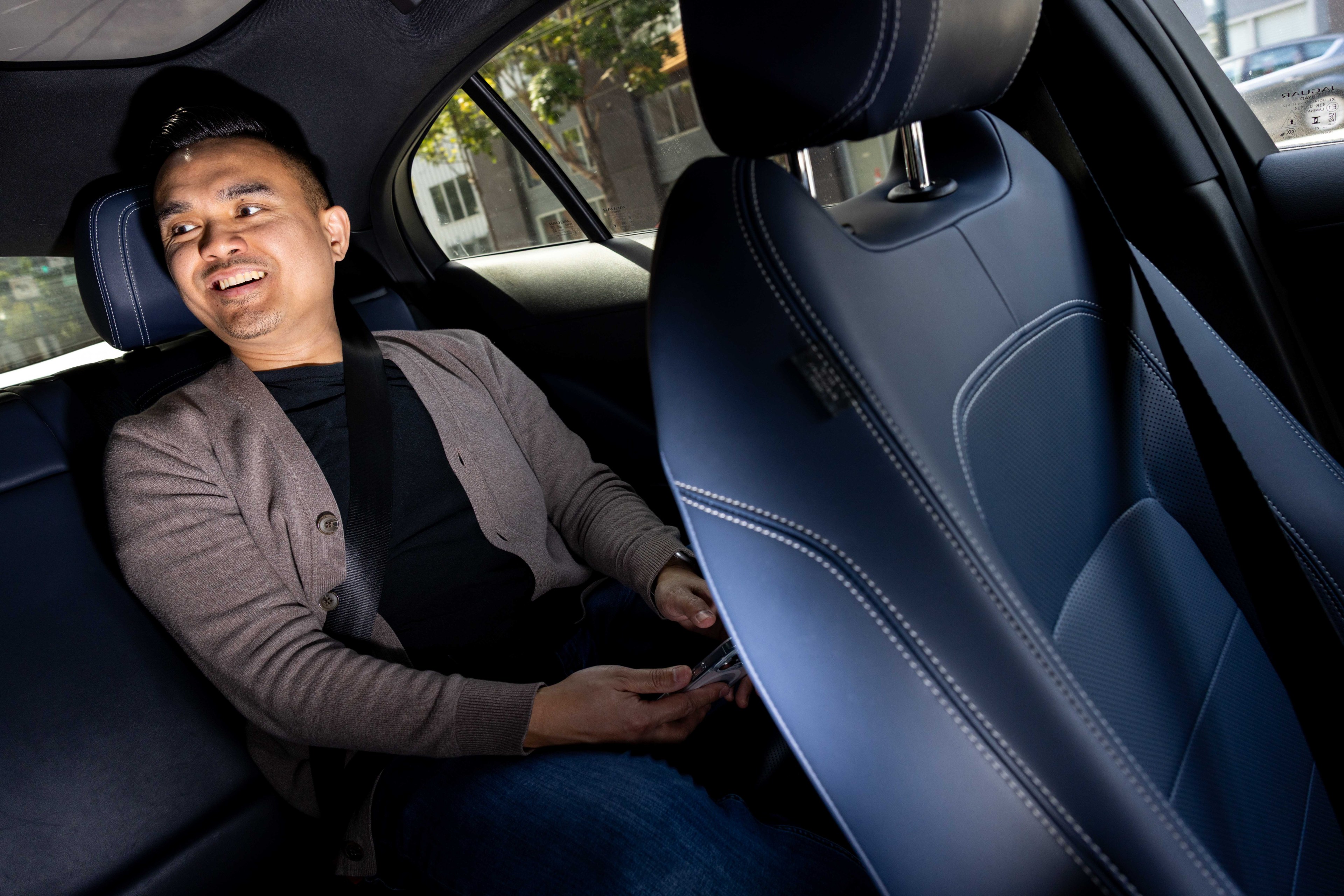 A person wearing a gray cardigan is smiling while seated in the backseat of a car, holding a phone. The car interior is dark with blue accents.