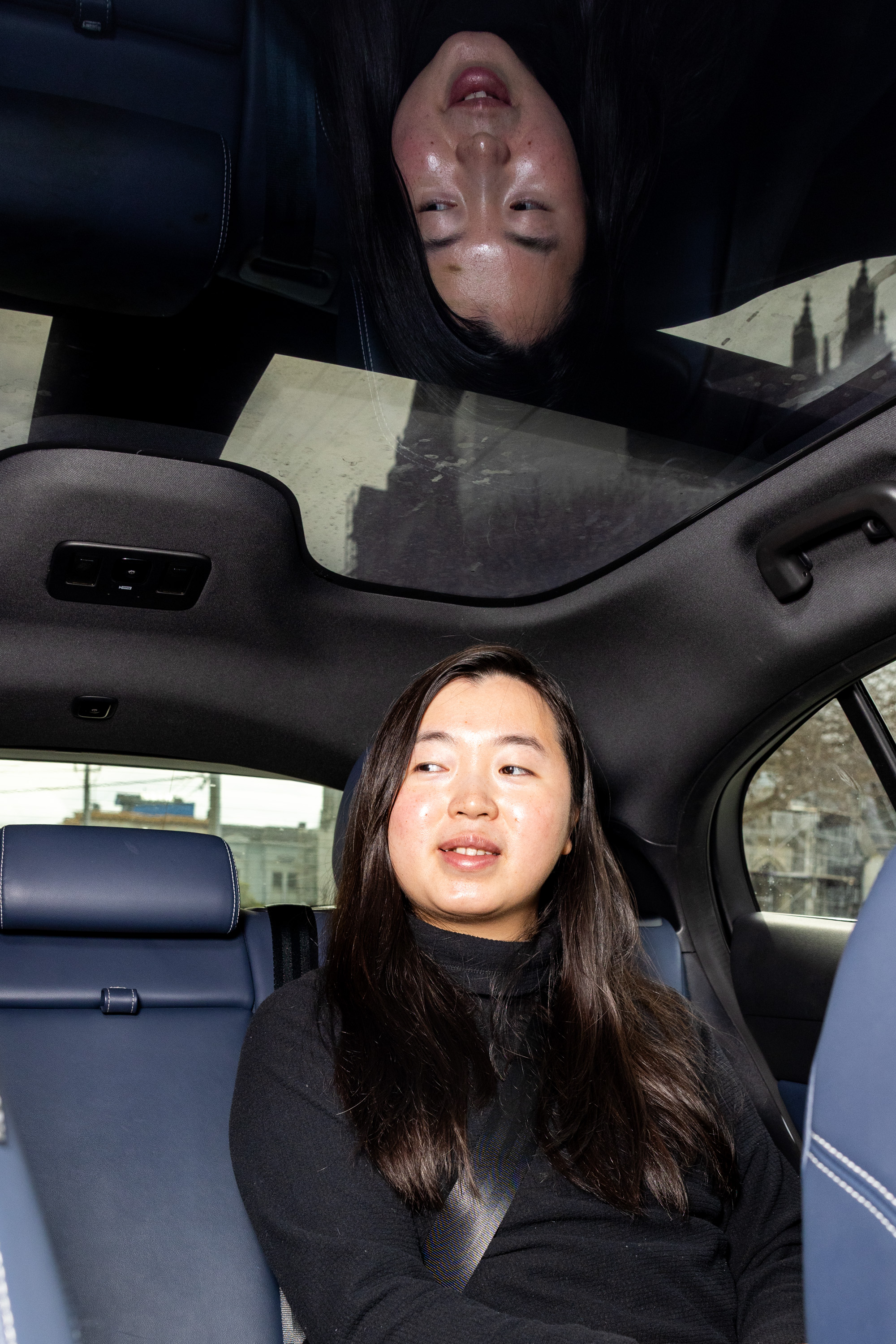 A person sits in a car's back seat, looking to the side. Their reflection is visible in the sunroof, and they are wearing a black sweater with a seatbelt fastened.