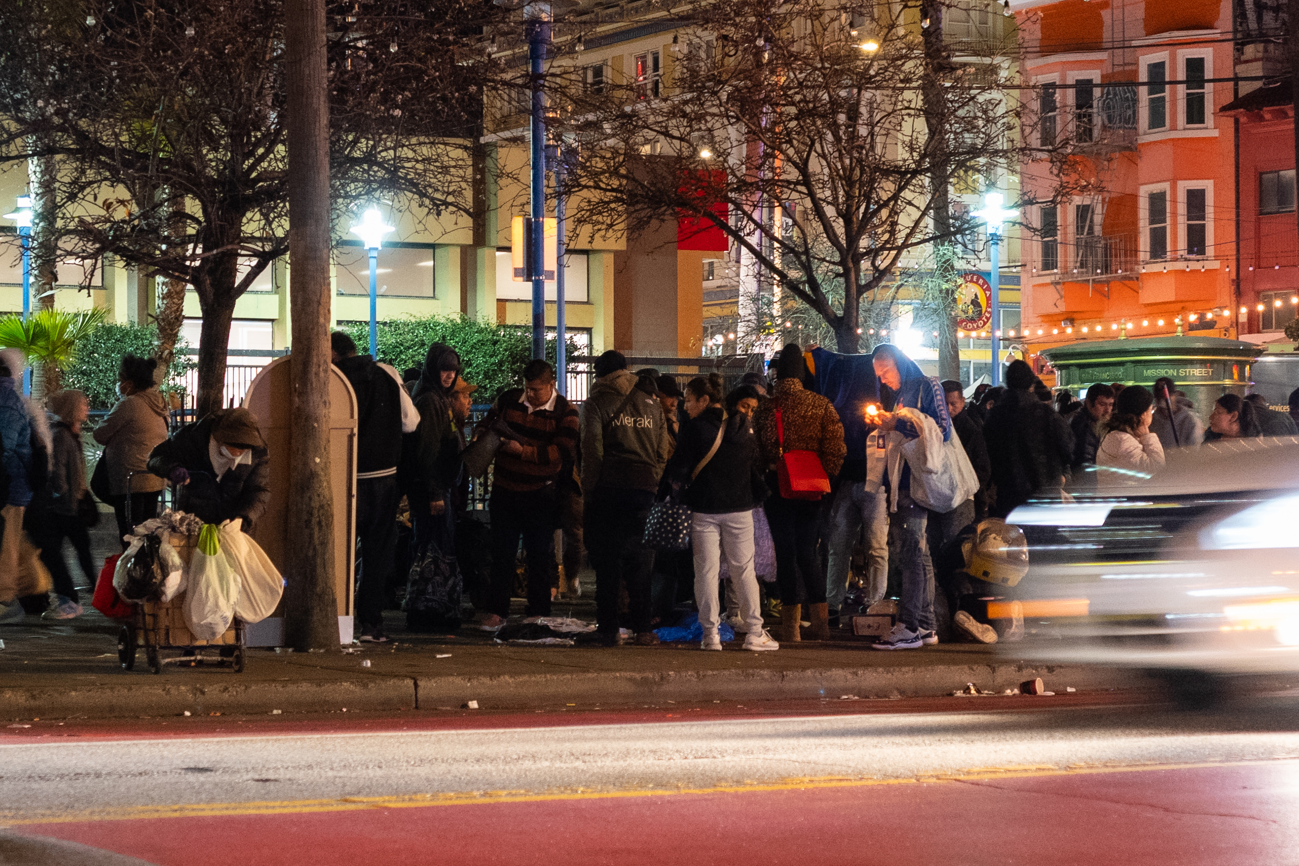 A group of people are gathered on a street at night, carrying bags and belongings. Some are looking down or conversing, with a lit candle visible.