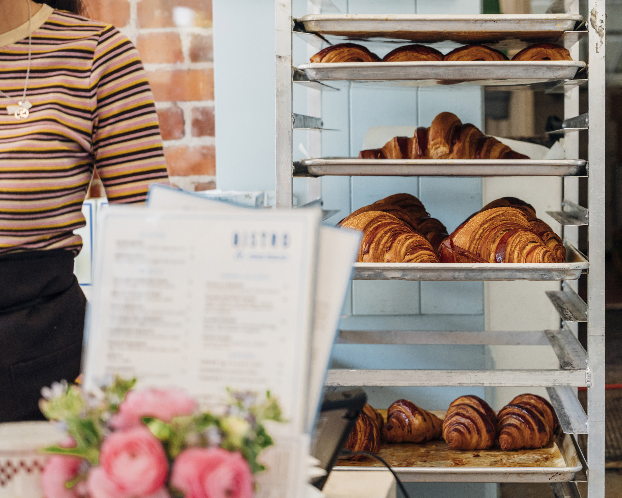 Racks hold trays of golden croissants in a bakery. A person in a striped shirt stands nearby, with menus and pink flowers in the foreground.