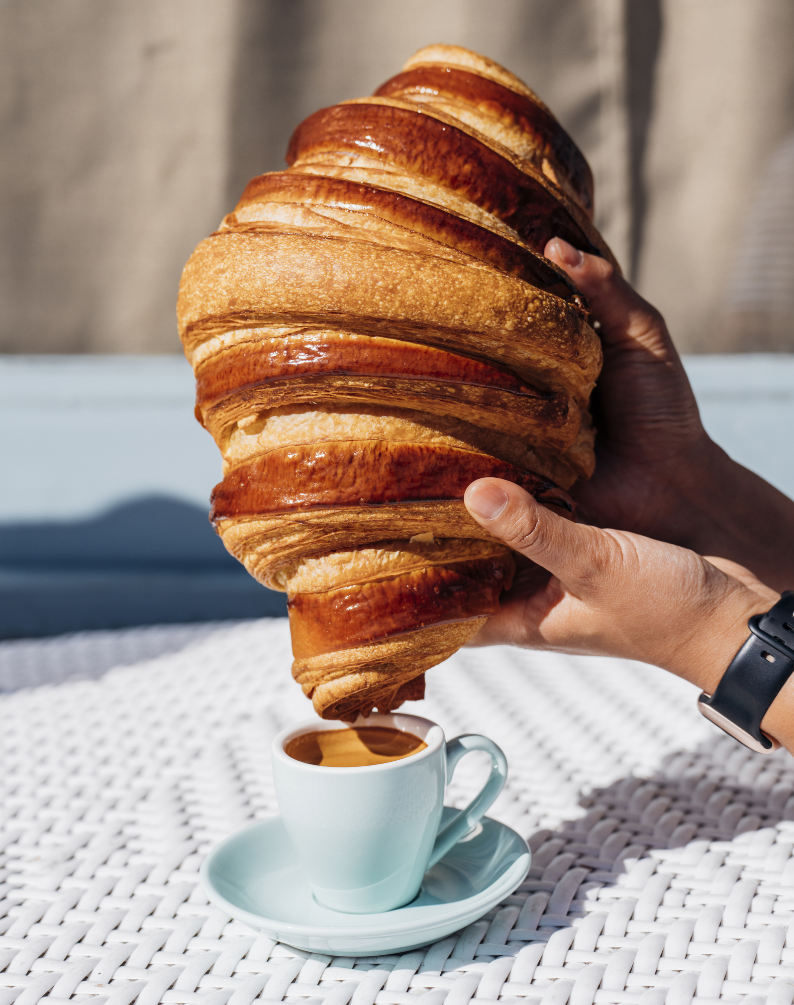 A person holds an oversized croissant above a small blue cup of coffee on a white textured table, creating a playful contrast in size.