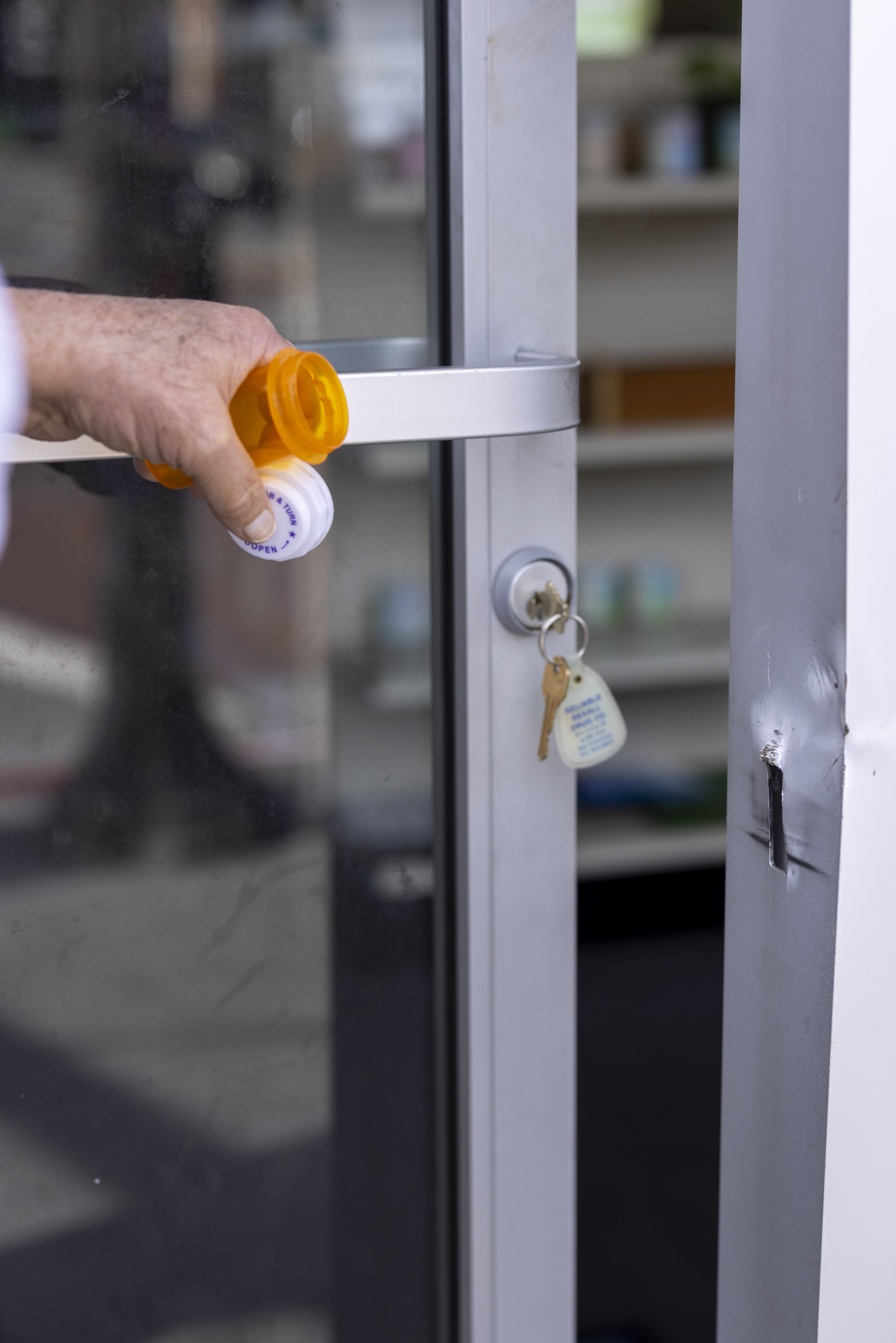 A hand holds an open orange pill bottle near a glass door. Keys with a tag hang from the lock, and a white shelf with bottles is visible in the background.