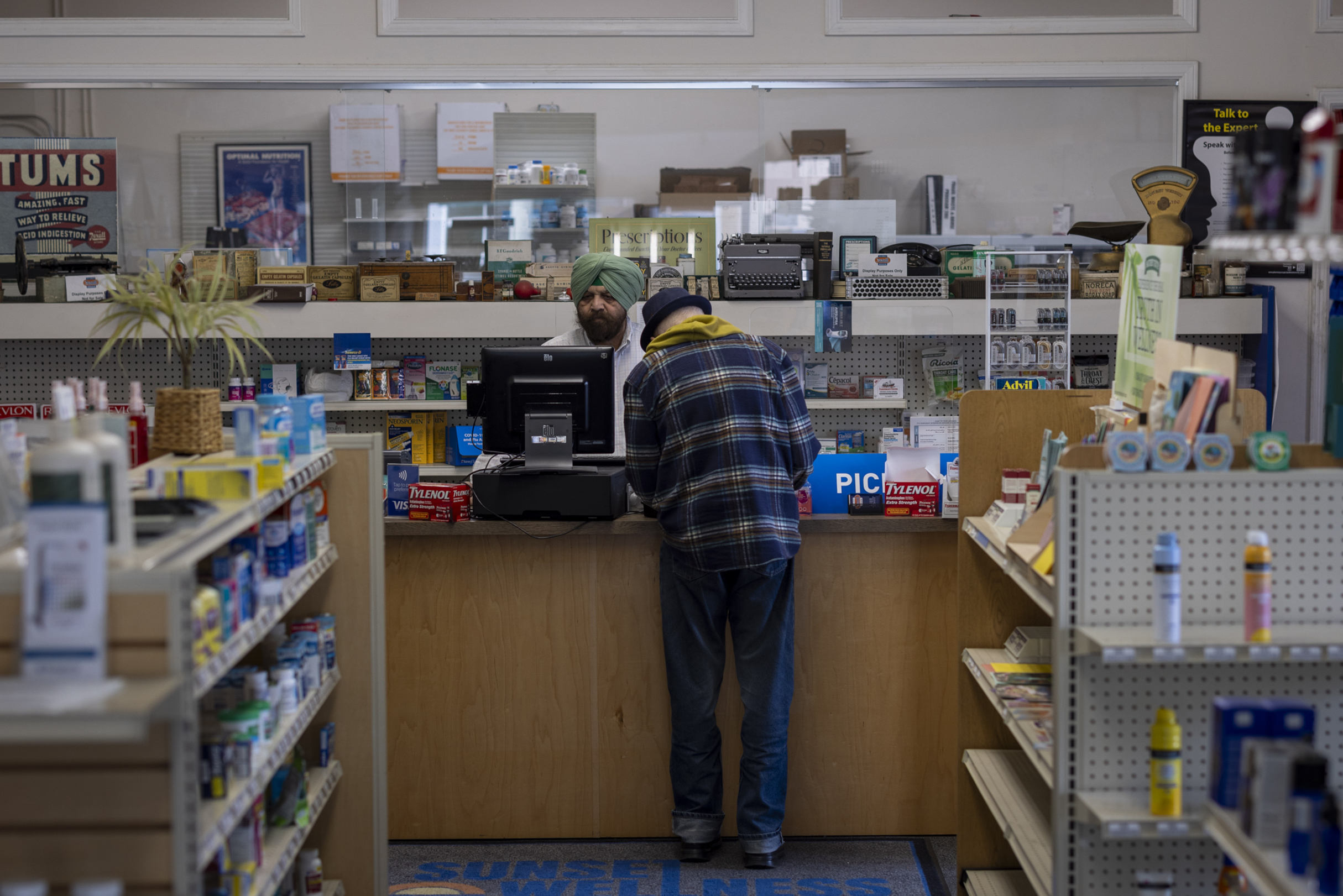 A man in a plaid jacket and beanie leans over a pharmacy counter talking to a staff member wearing a green turban behind the register, surrounded by shelves of goods.