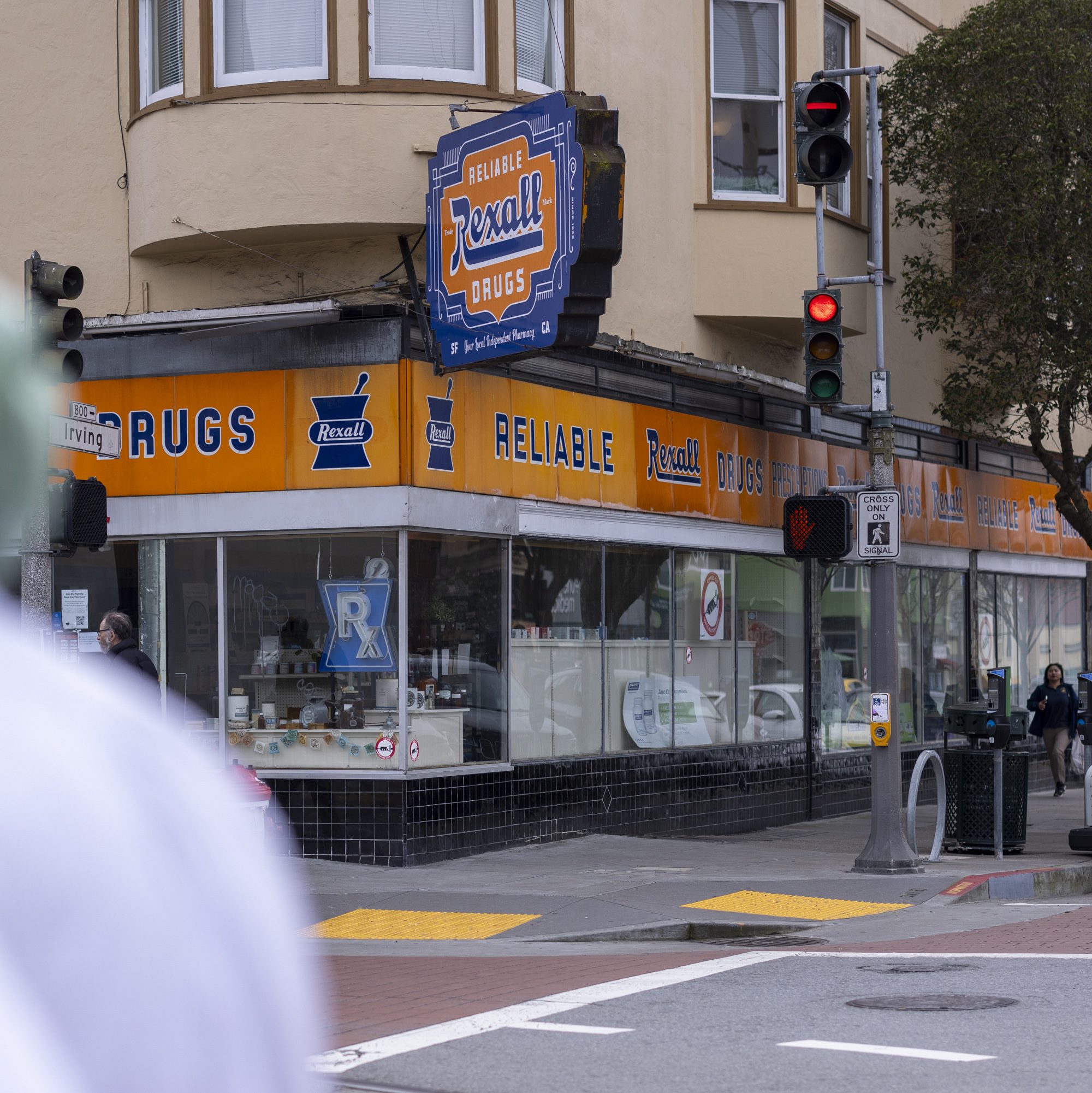 The image shows a street corner with a &quot;Reliable Rexall Drugs&quot; store, featuring a retro sign and orange facade. A traffic light is red, and pedestrians are nearby.