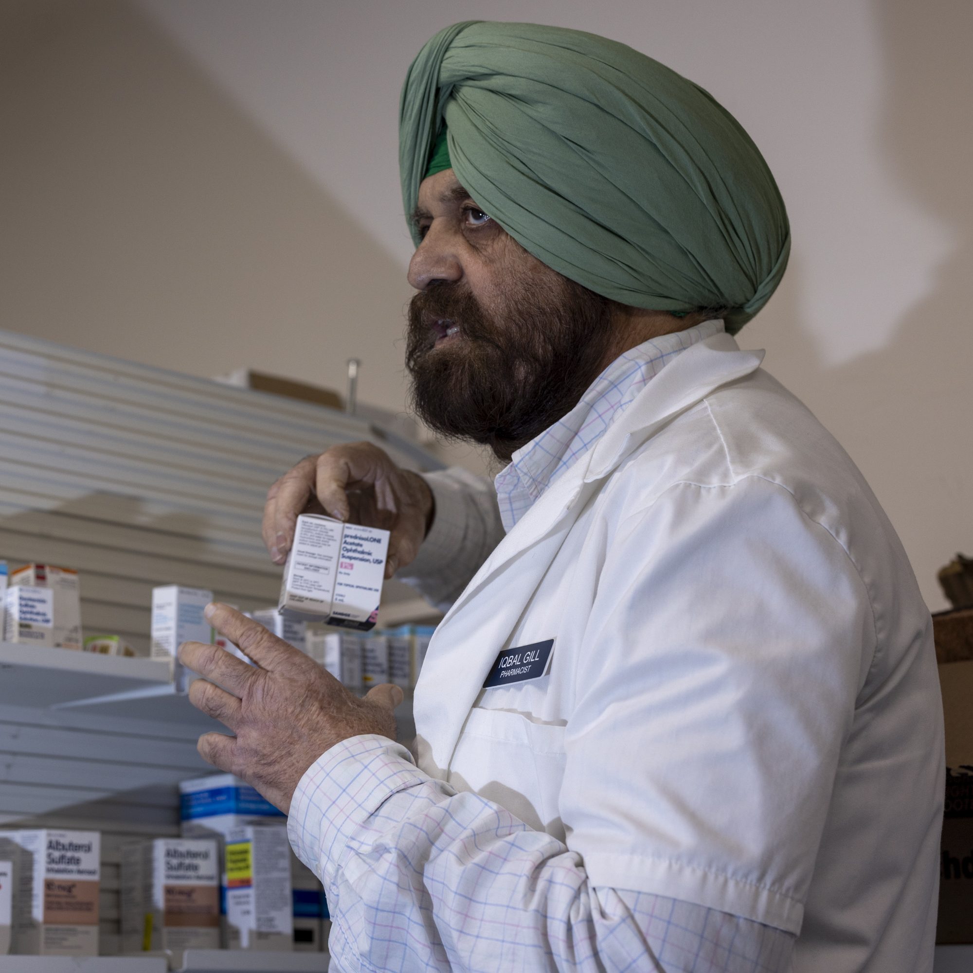 A pharmacist in a lab coat and green turban is holding a box of medication, standing in front of a shelf with various medicine boxes.