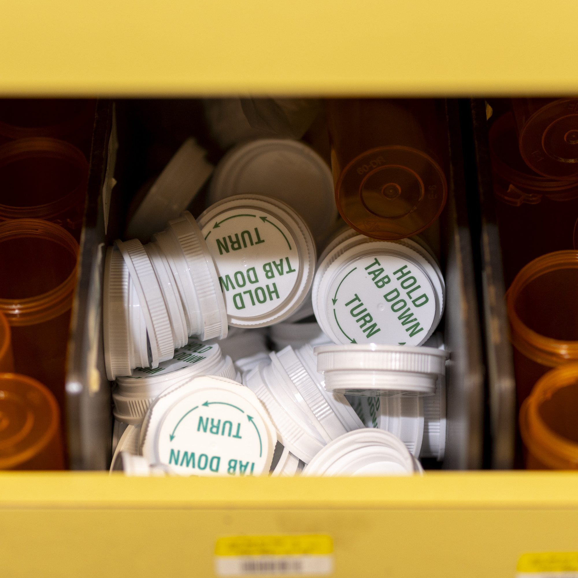 The image shows a drawer filled with white plastic medicine bottle caps, labeled &quot;HOLD TAB DOWN TURN&quot; in green. Orange bottle openings are visible around them.