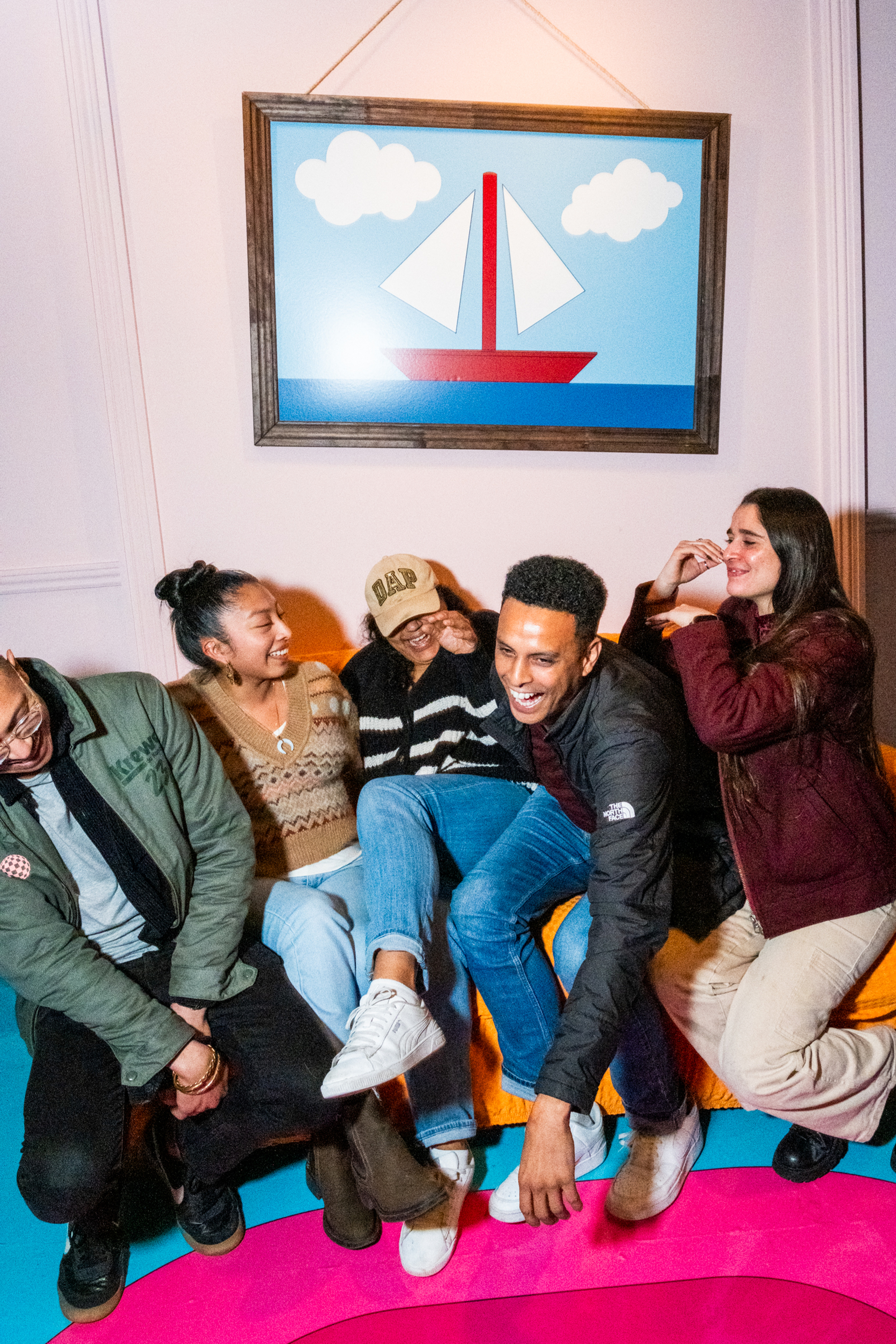 Five people are sitting closely, laughing and enjoying each other's company. A colorful painting of a sailboat is hanging on the wall behind them.