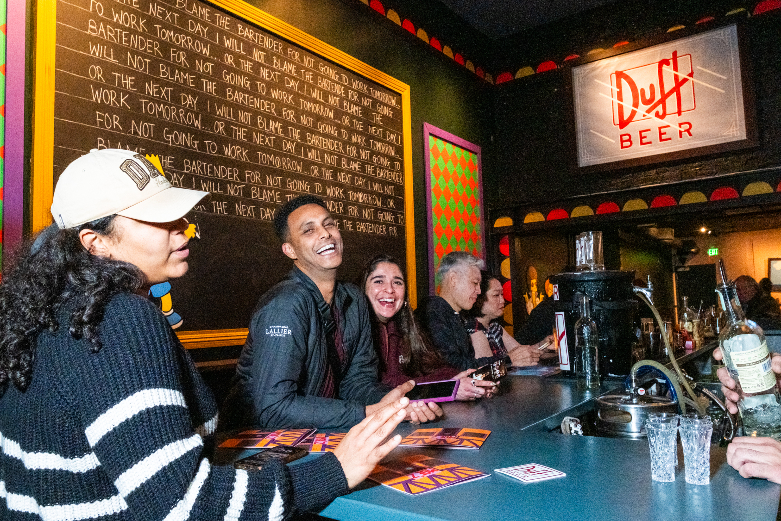 A group of people are laughing and chatting at a colorful bar with a "Duff Beer" sign. The atmosphere is lively, and drinks are being served.