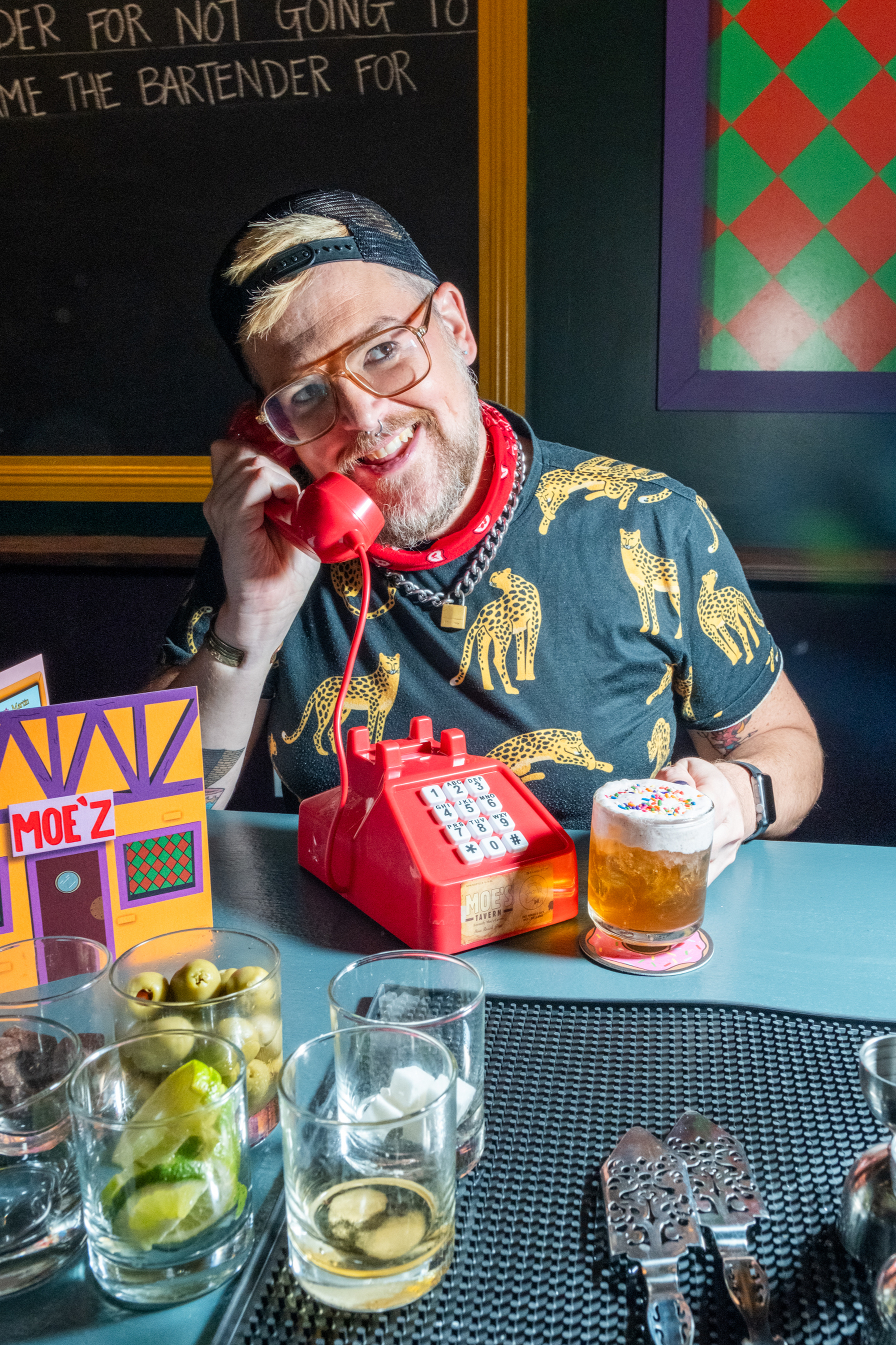 A smiling person in glasses and a giraffe-print shirt holds a red phone at a bar with a drink topped with whipped cream and sprinkles.