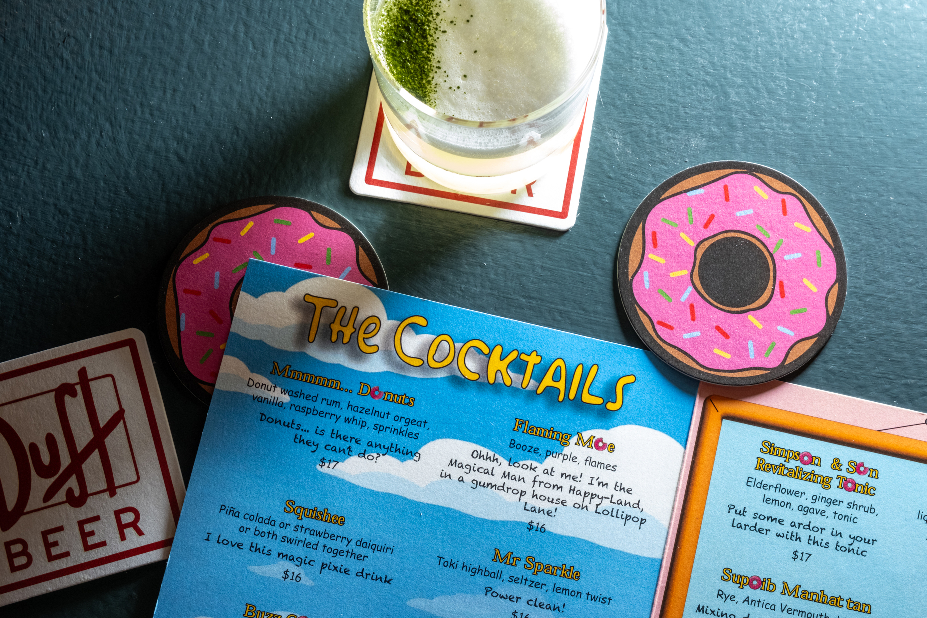 A cocktail menu featuring playful drink names is surrounded by donut-shaped coasters and a glass of a frothy green drink on a &quot;Duff Beer&quot; coaster.