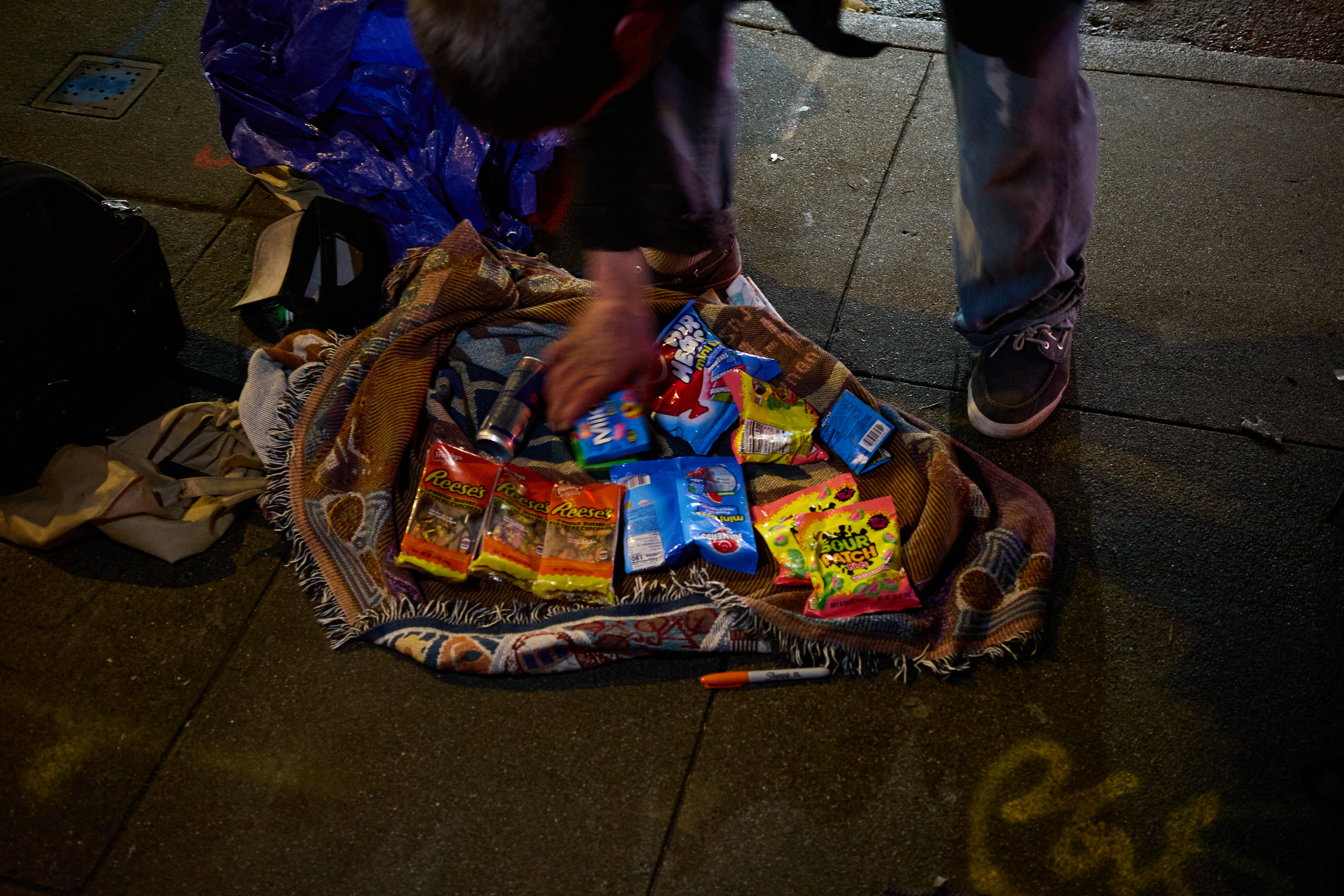 A person arranges snacks and candies on a blanket laid on the sidewalk, including Reese's and Sour Patch Kids. A marker and bags are nearby.