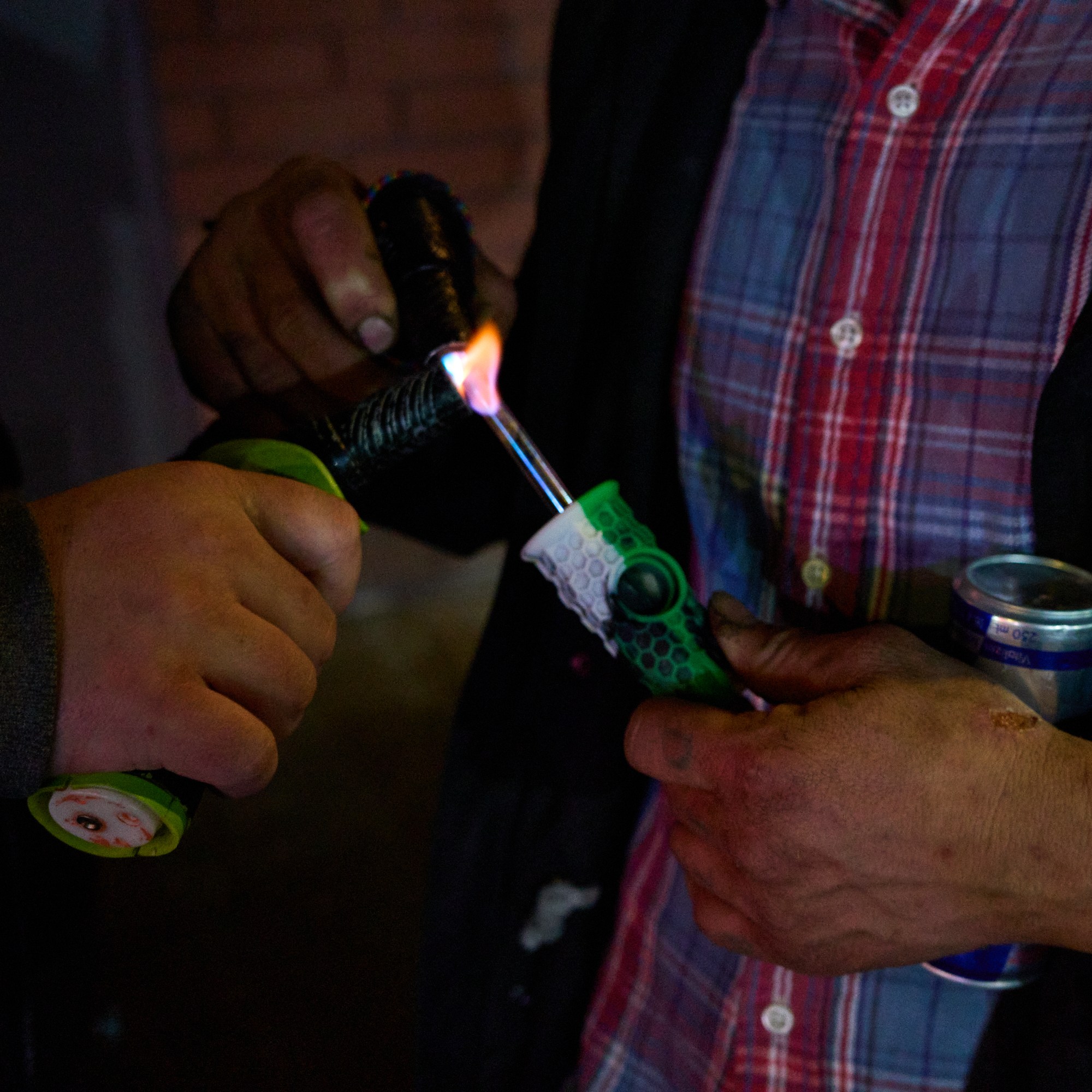 Two people are using a torch lighter to heat a pipe. One is holding a can, and the other is wearing a red plaid shirt and a dark jacket.