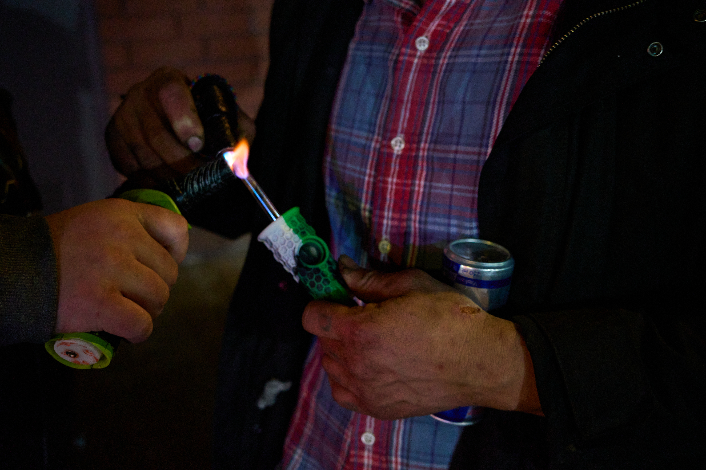 Two people are using a torch lighter to heat a pipe. One is holding a can, and the other is wearing a red plaid shirt and a dark jacket.