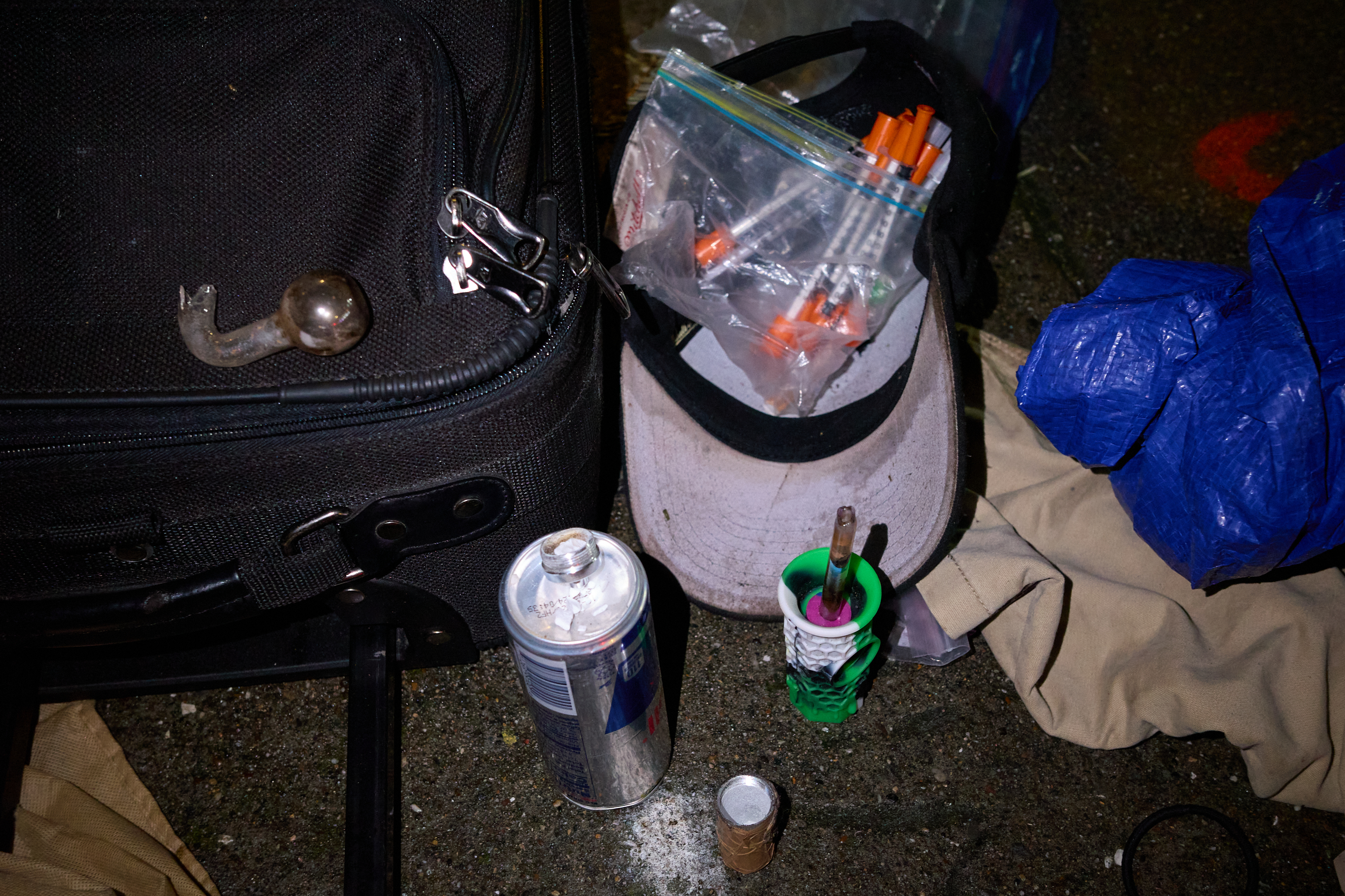 The image shows a suitcase, a can, a cap filled with plastic bags of syringes, a small container with a pen, and blue and beige fabrics on the ground.