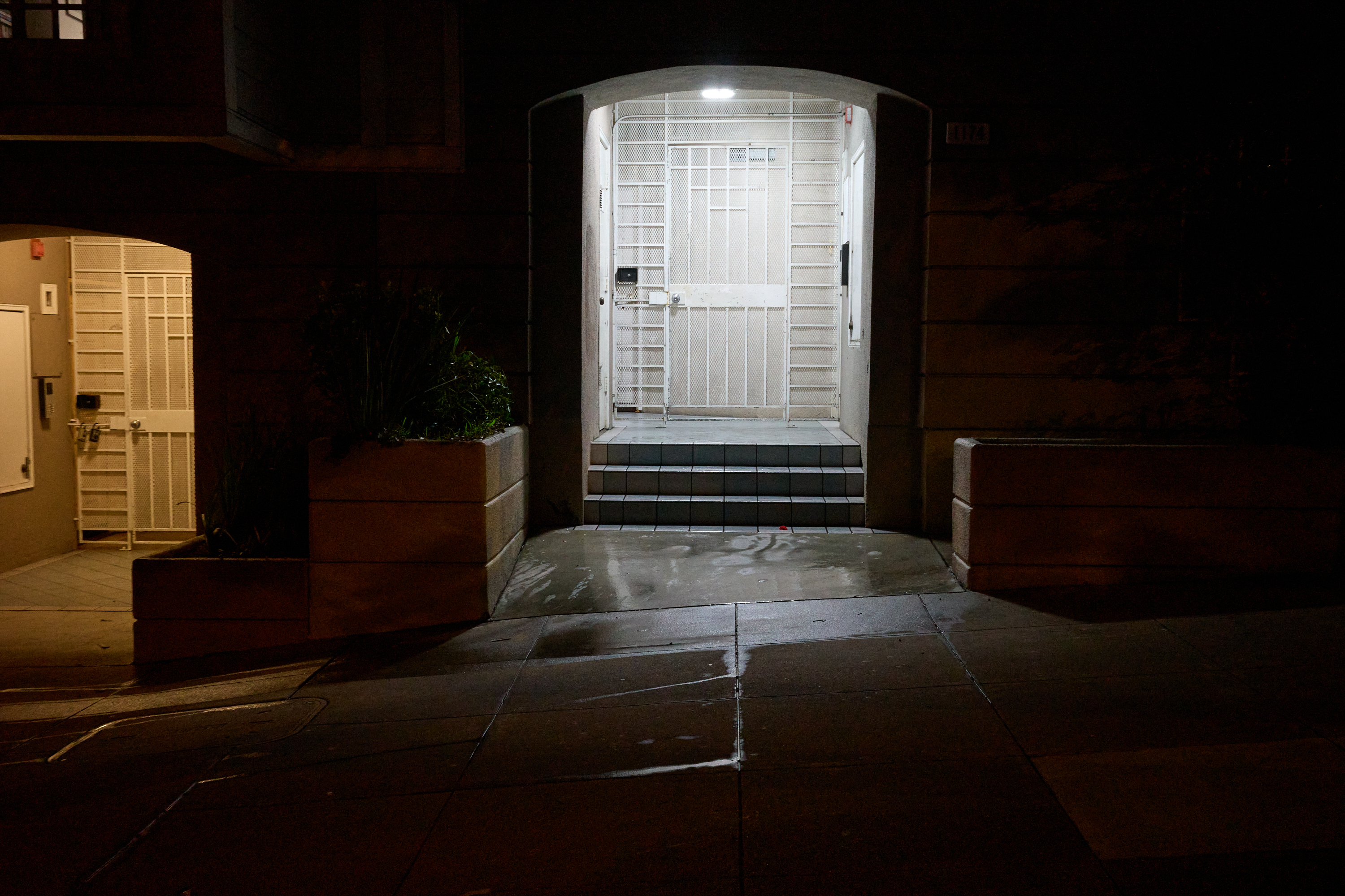 A dimly lit entrance with a metal gate and light overhead. It has tiled steps, flanked by large plant containers, leading from a wet sidewalk.