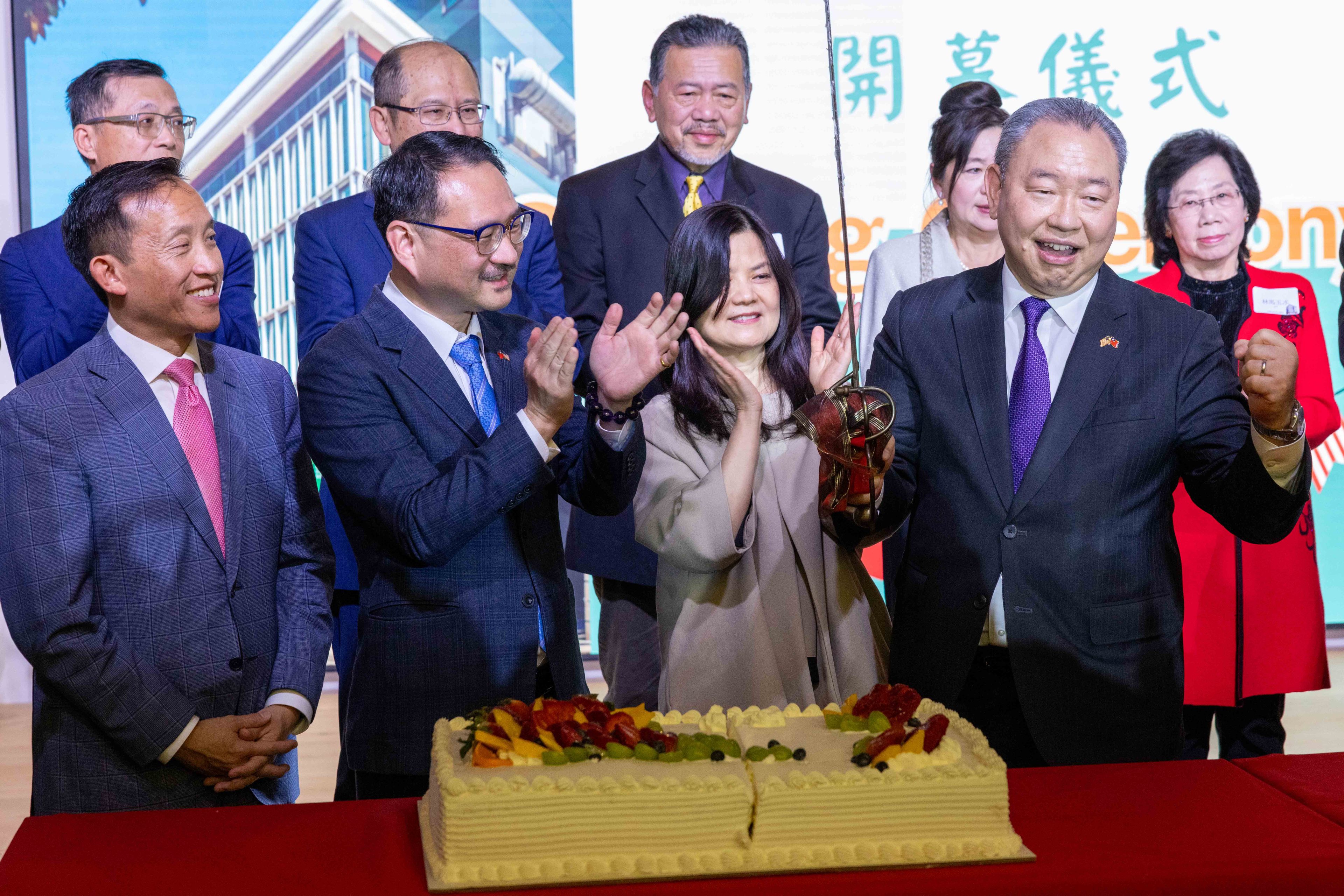 A group of people is celebrating in front of a cake with fruit decorations. They appear happy and are clapping, with one person holding scissors.