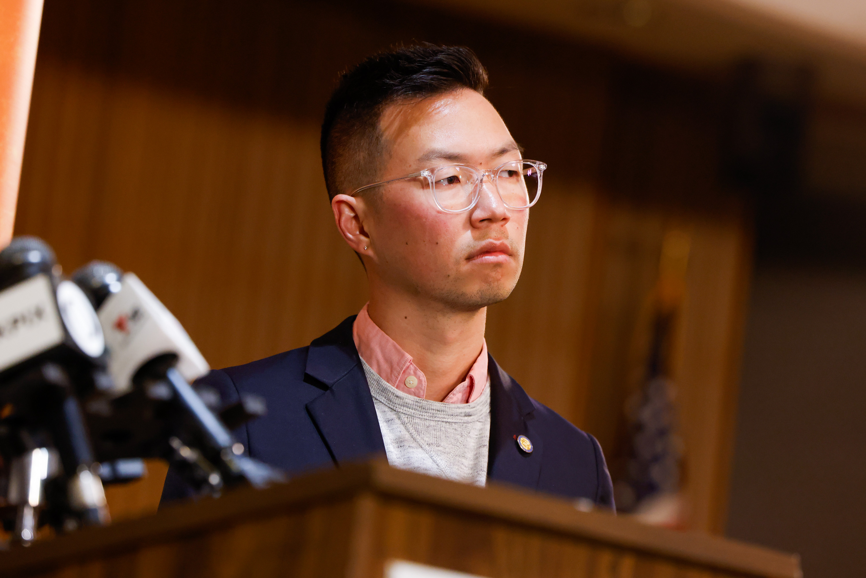 A person with short hair and glasses stands behind a podium with microphones, wearing a suit and a pink shirt, looking serious. An American flag is blurred in the background.