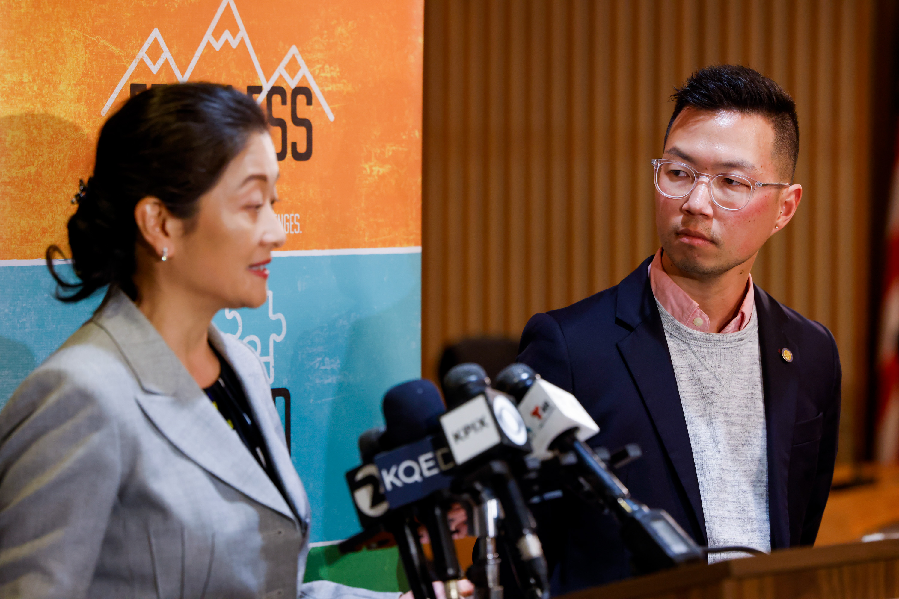 Two people stand at a podium with microphones. The woman speaks, while the man looks on. A colorful banner with text and images is behind them.