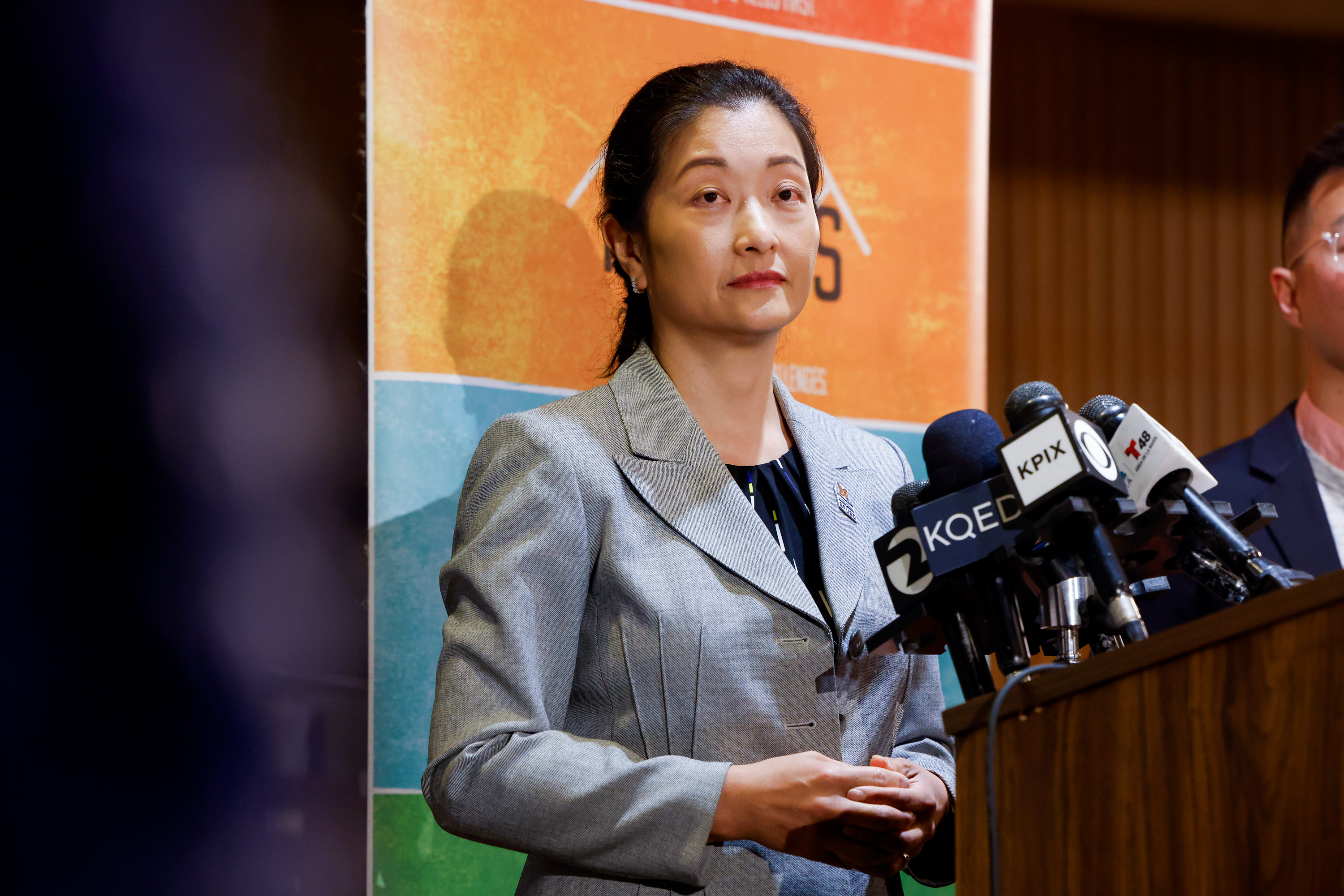 A woman in a gray blazer speaks at a press conference. She stands in front of a colorful backdrop with multiple microphones labeled with news stations.