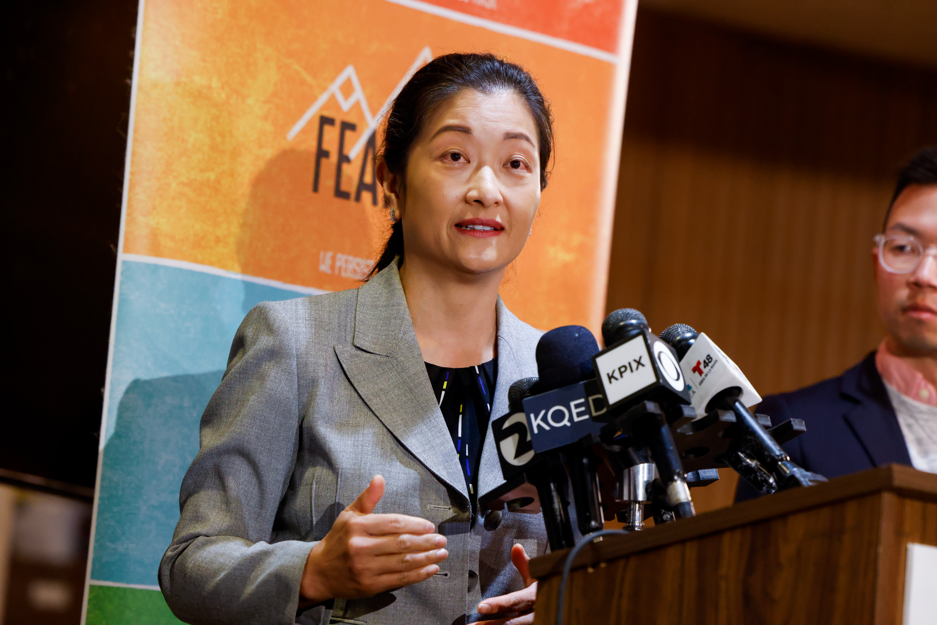 A person in a gray jacket speaks at a podium with multiple microphones. A colorful banner is visible in the background, and another person stands nearby.