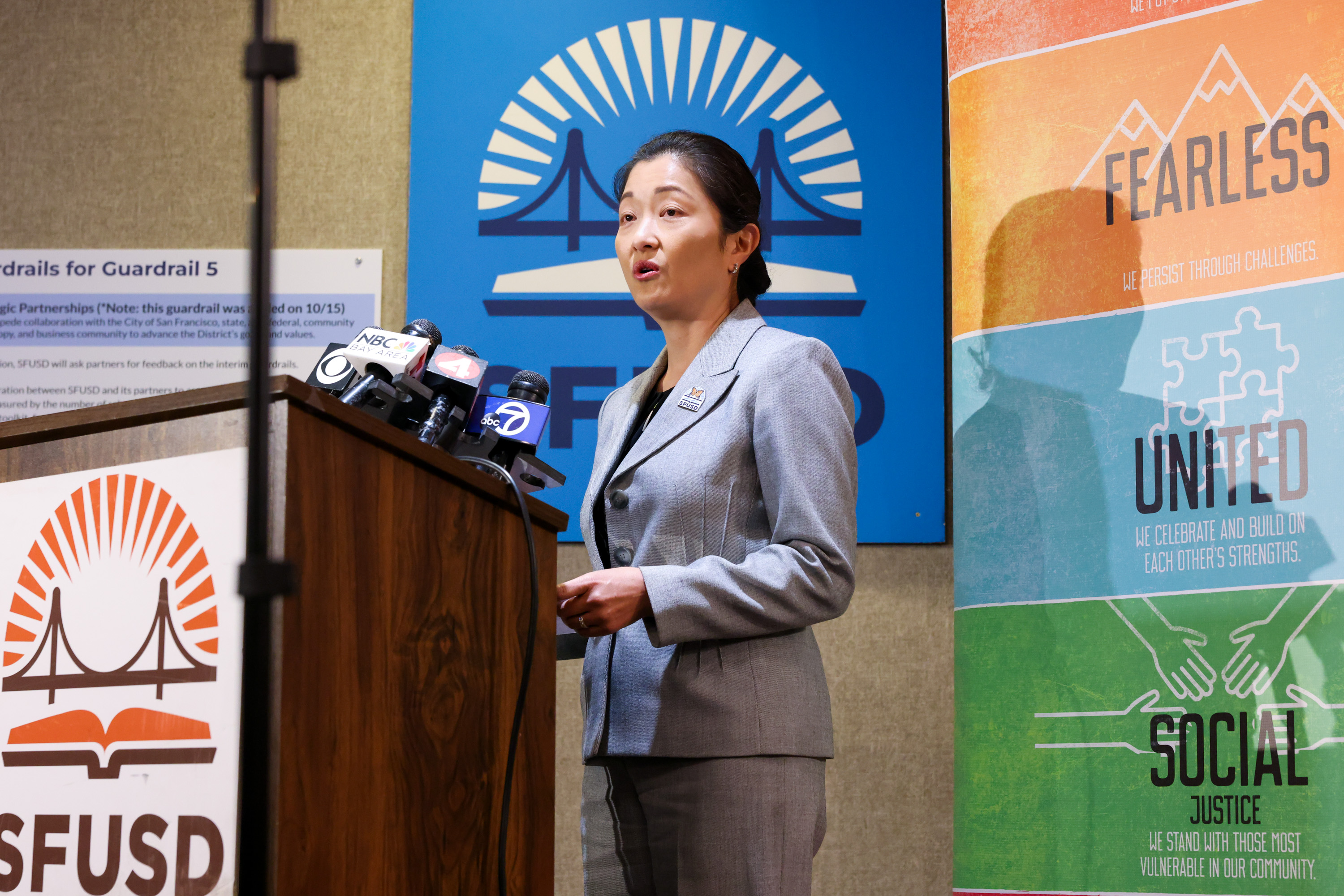 A person in a gray suit speaks at a podium with multiple microphones. Behind are banners with text like &quot;FEARLESS,&quot; &quot;UNITED,&quot; and &quot;SOCIAL JUSTICE.&quot;
