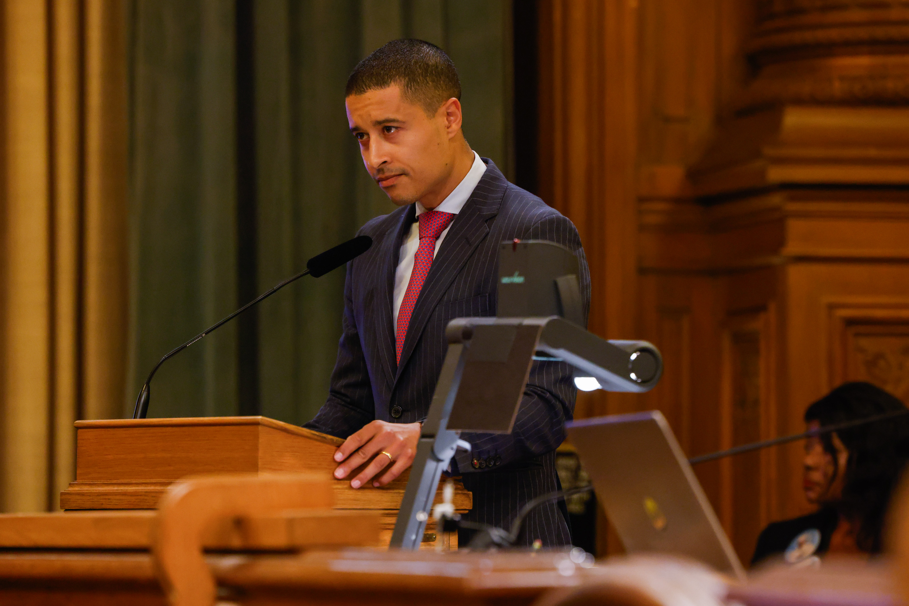 A person in a suit speaks at a wooden podium with a microphone. The setting appears formal, likely a conference or meeting room, with a laptop nearby.