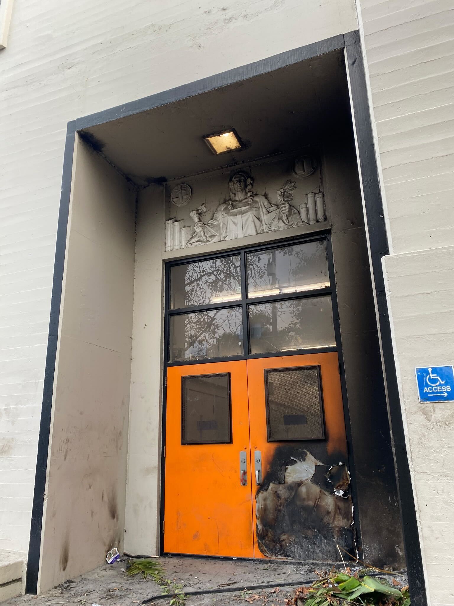 A pair of orange doors with visible fire damage at the bottom is set into a gray building entrance. Above is a decorative relief, and a handicap access sign is on the side.