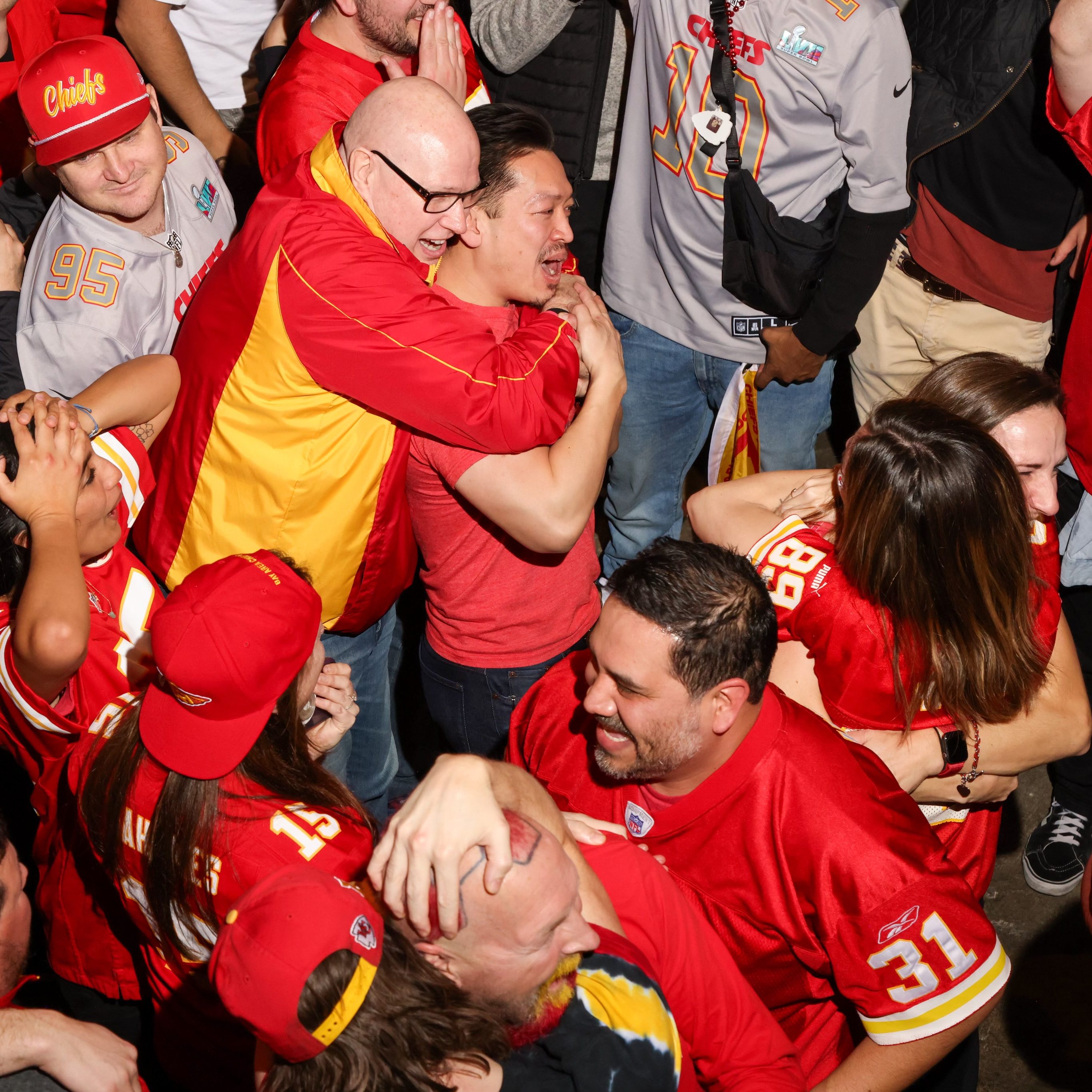 A group of people in red and yellow Kansas City Chiefs gear are joyfully hugging and celebrating. They seem excited, with big smiles and some raised arms.