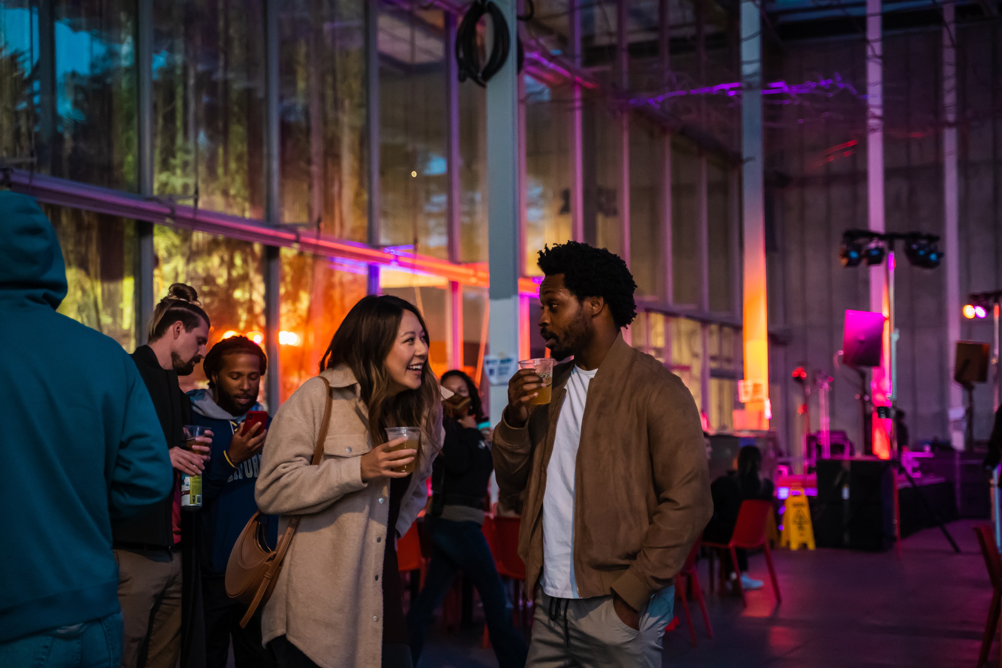 A lively gathering in a dimly lit space with colorful lights. People are talking and holding drinks, creating a relaxed and social atmosphere.