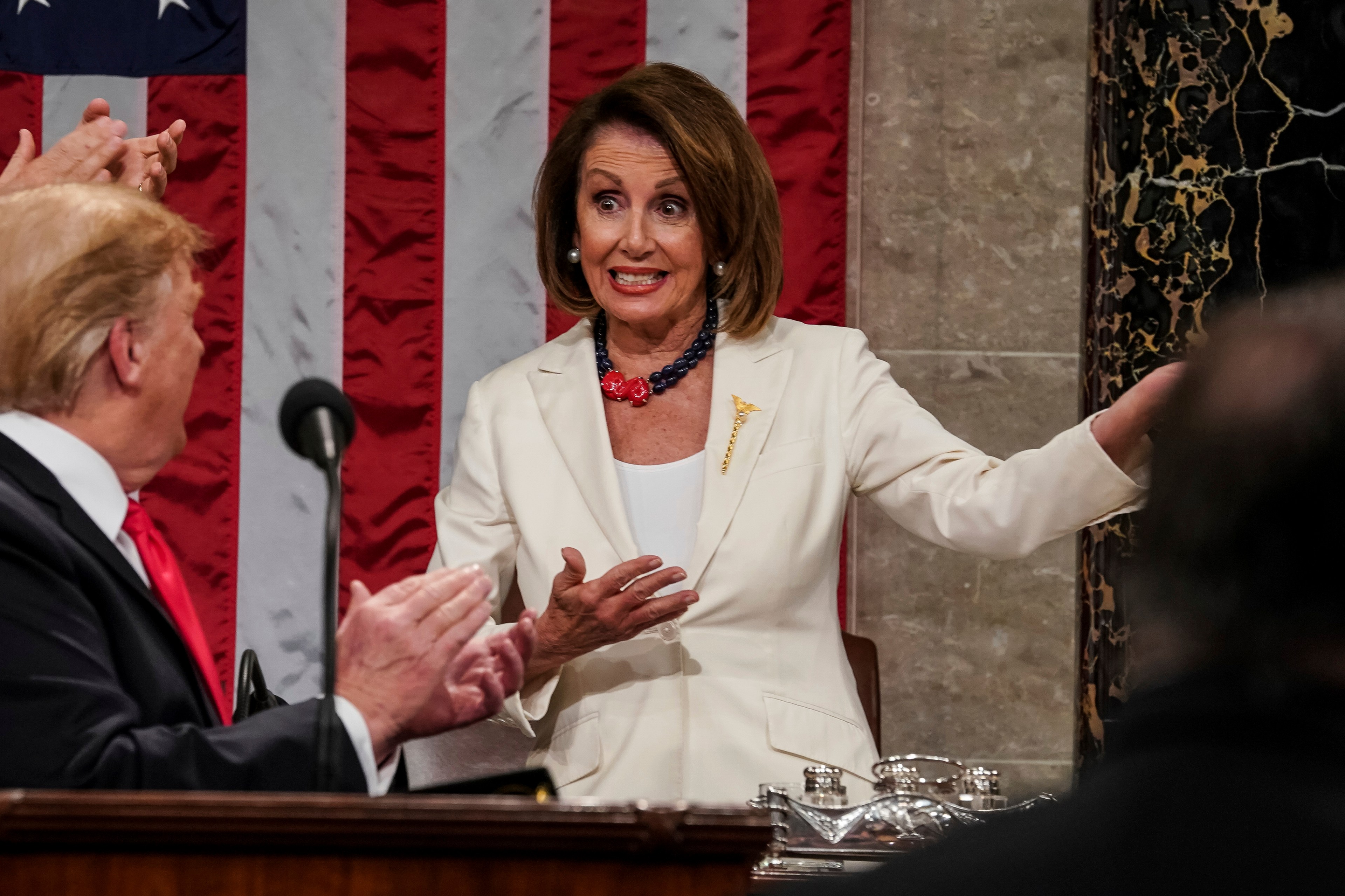 A woman in a white suit gestures with her hand while a man claps in the foreground. An American flag hangs in the background, and others observe.