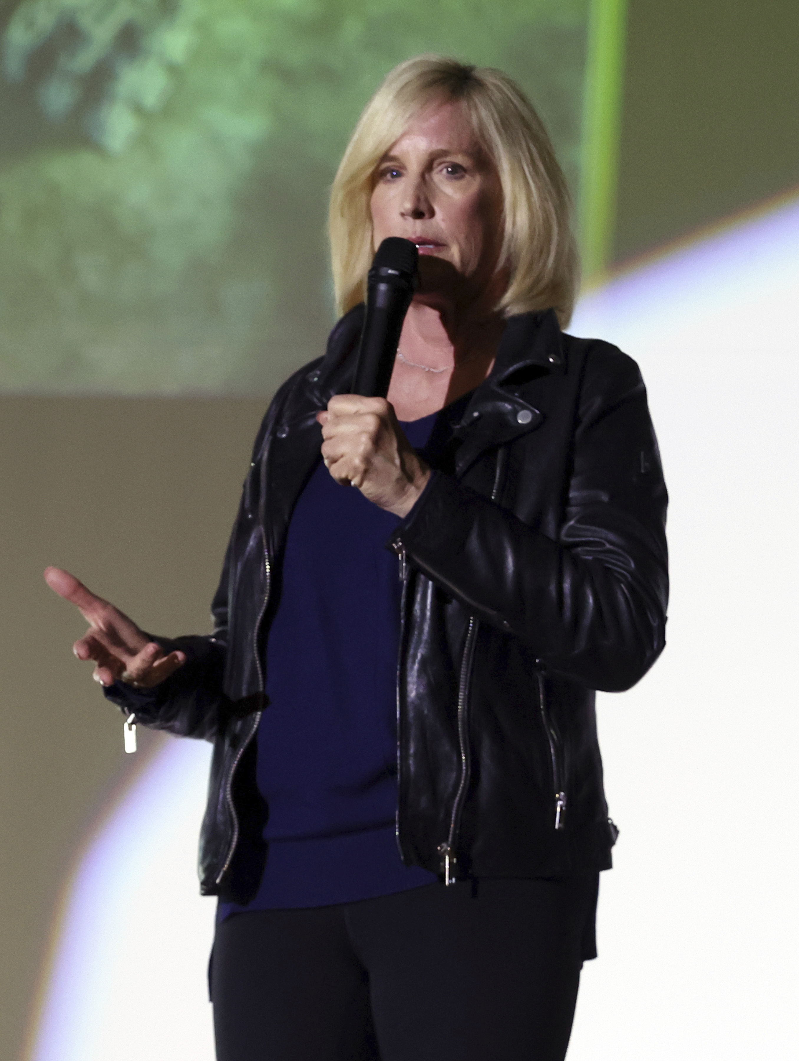 A person with blonde hair is speaking into a microphone. They are wearing a black leather jacket and standing against a blurred background.