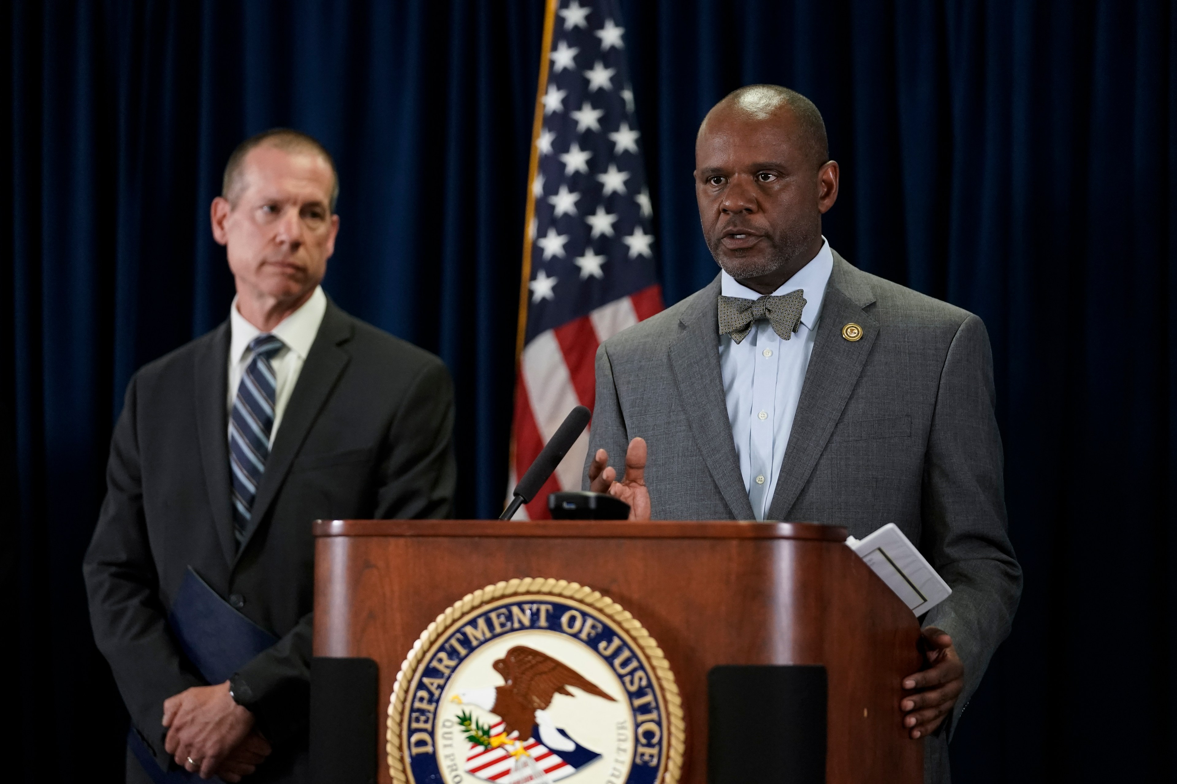 Two men in suits stand at a podium with the Department of Justice emblem. The man on the right speaks, while the one on the left listens. An American flag is behind them.