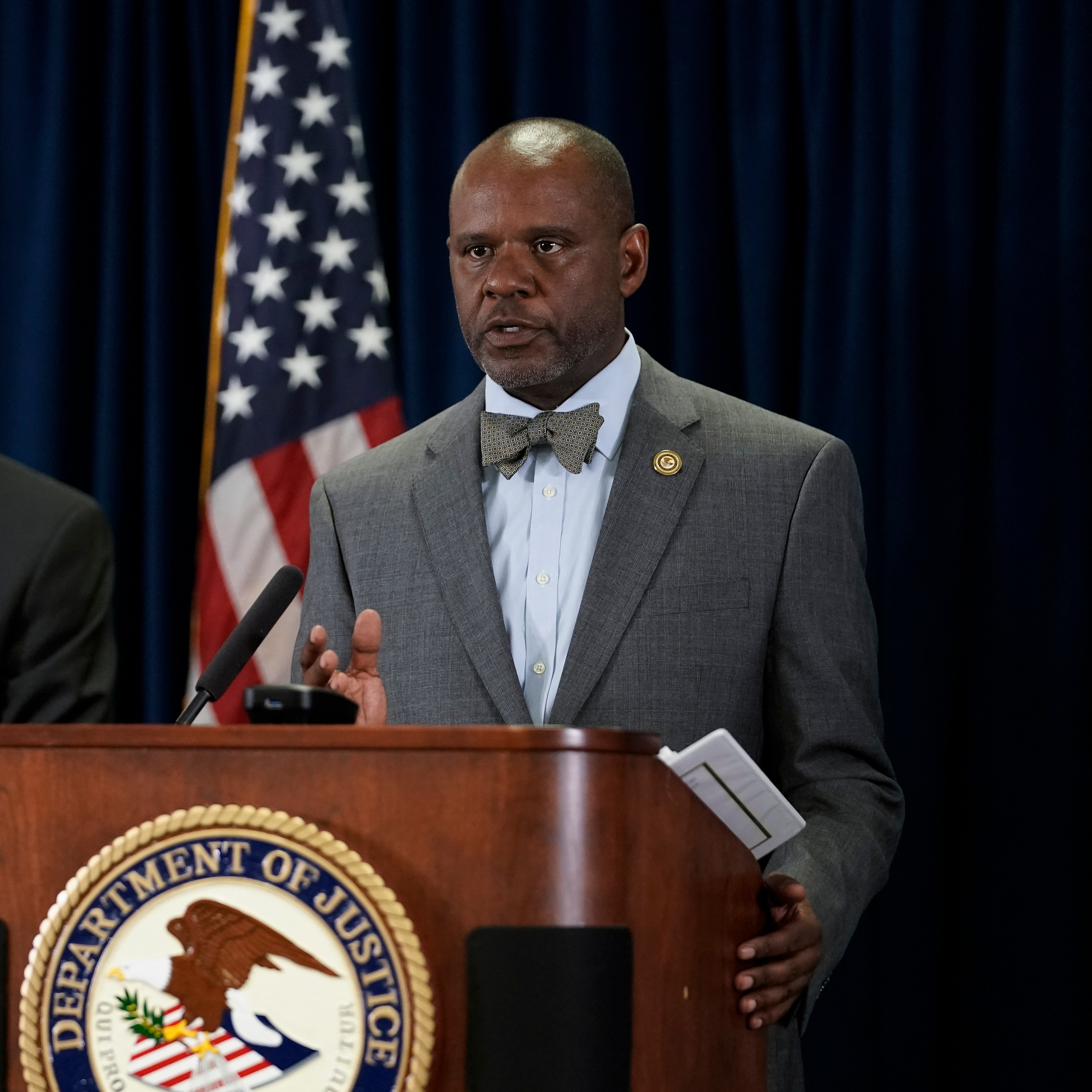 Two men in suits stand at a podium with the Department of Justice emblem. The man on the right speaks, while the one on the left listens. An American flag is behind them.