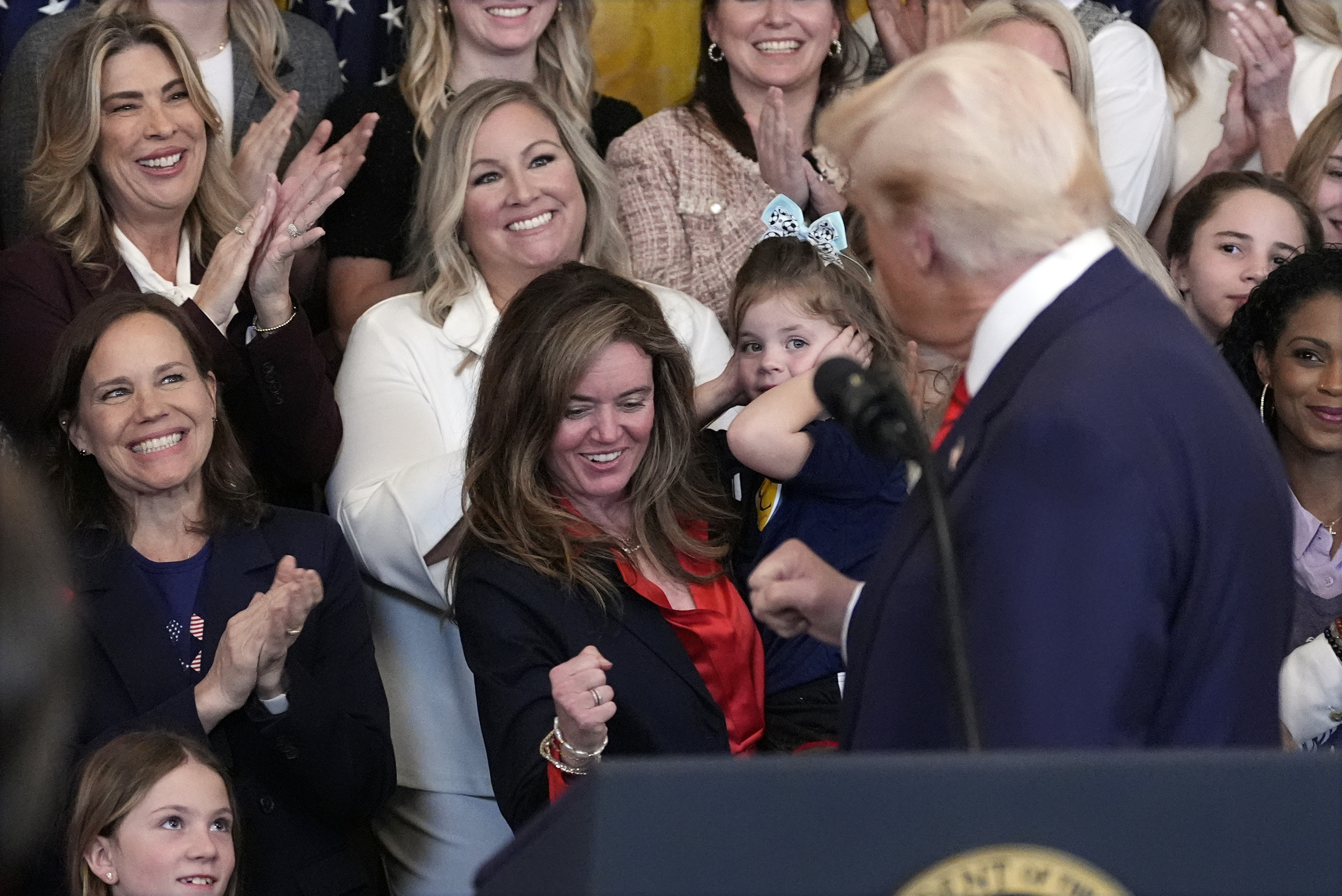 A group of people, including women and a child, are gathered together, smiling and clapping. A man facing them speaks from a podium.