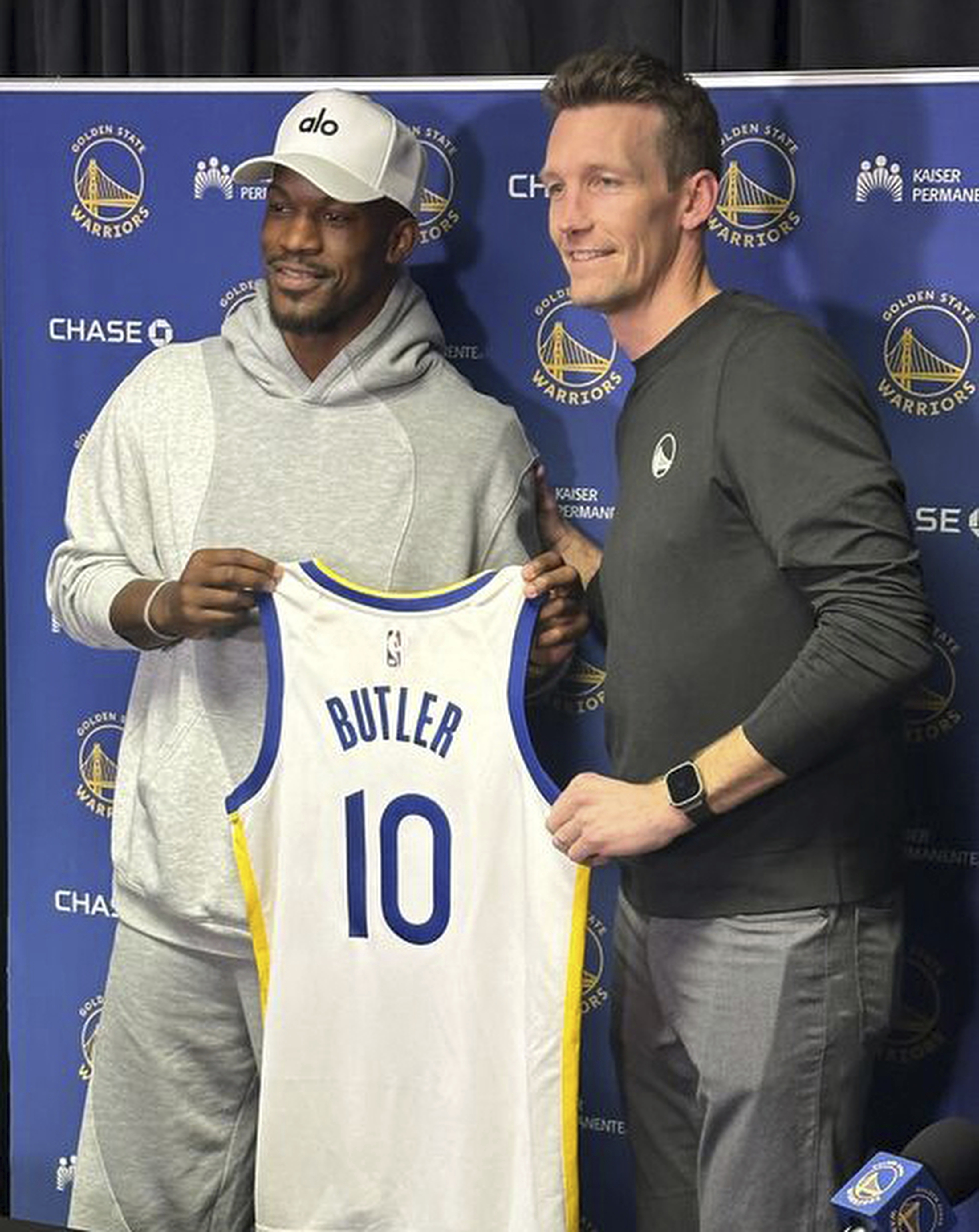 Two people stand together smiling, one holding a Golden State Warriors jersey with &quot;Butler&quot; and number 10. They're in front of a branded backdrop.