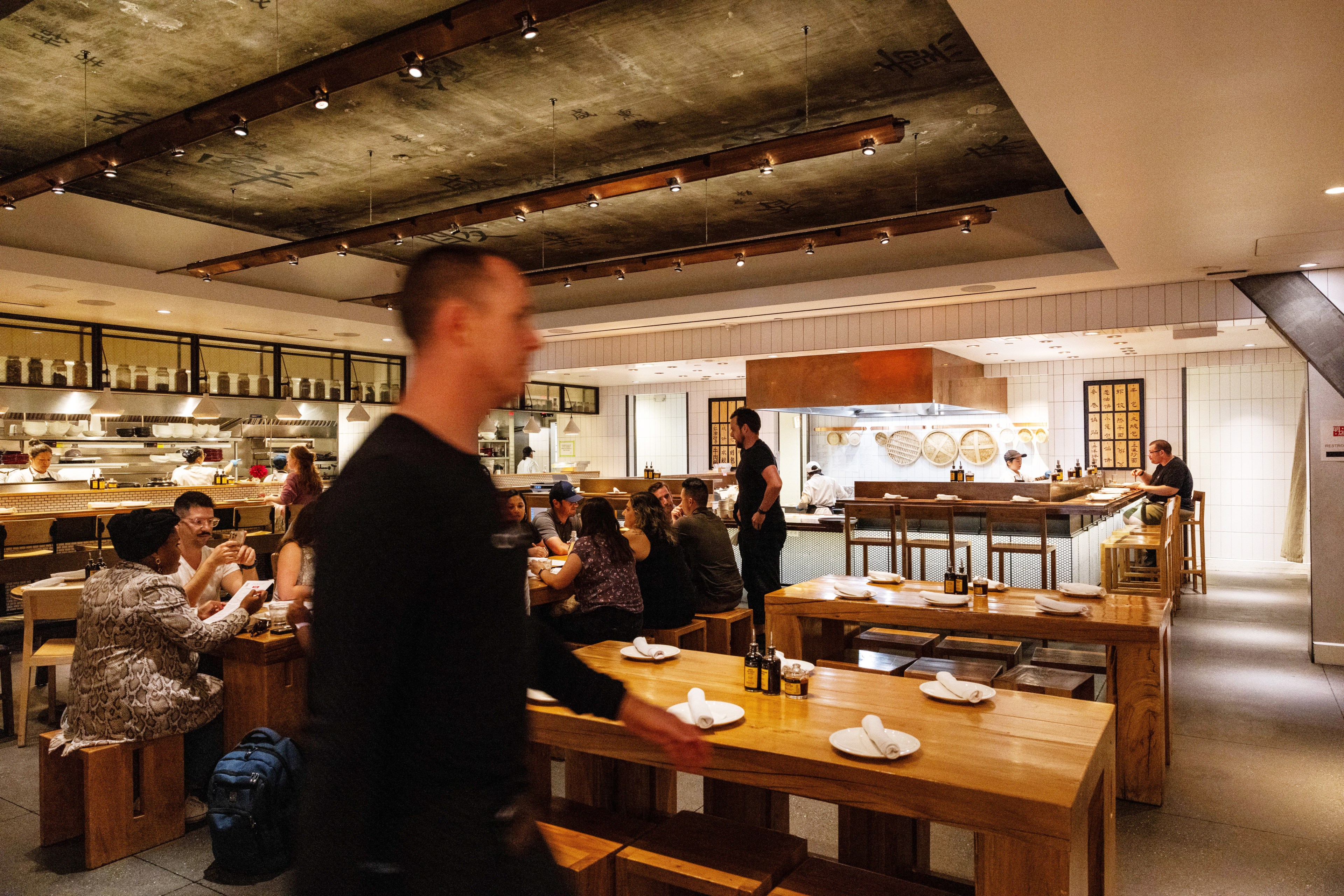 A bustling restaurant with wooden tables, diners chatting, and staff in motion. An open kitchen is in the background with chefs preparing food.