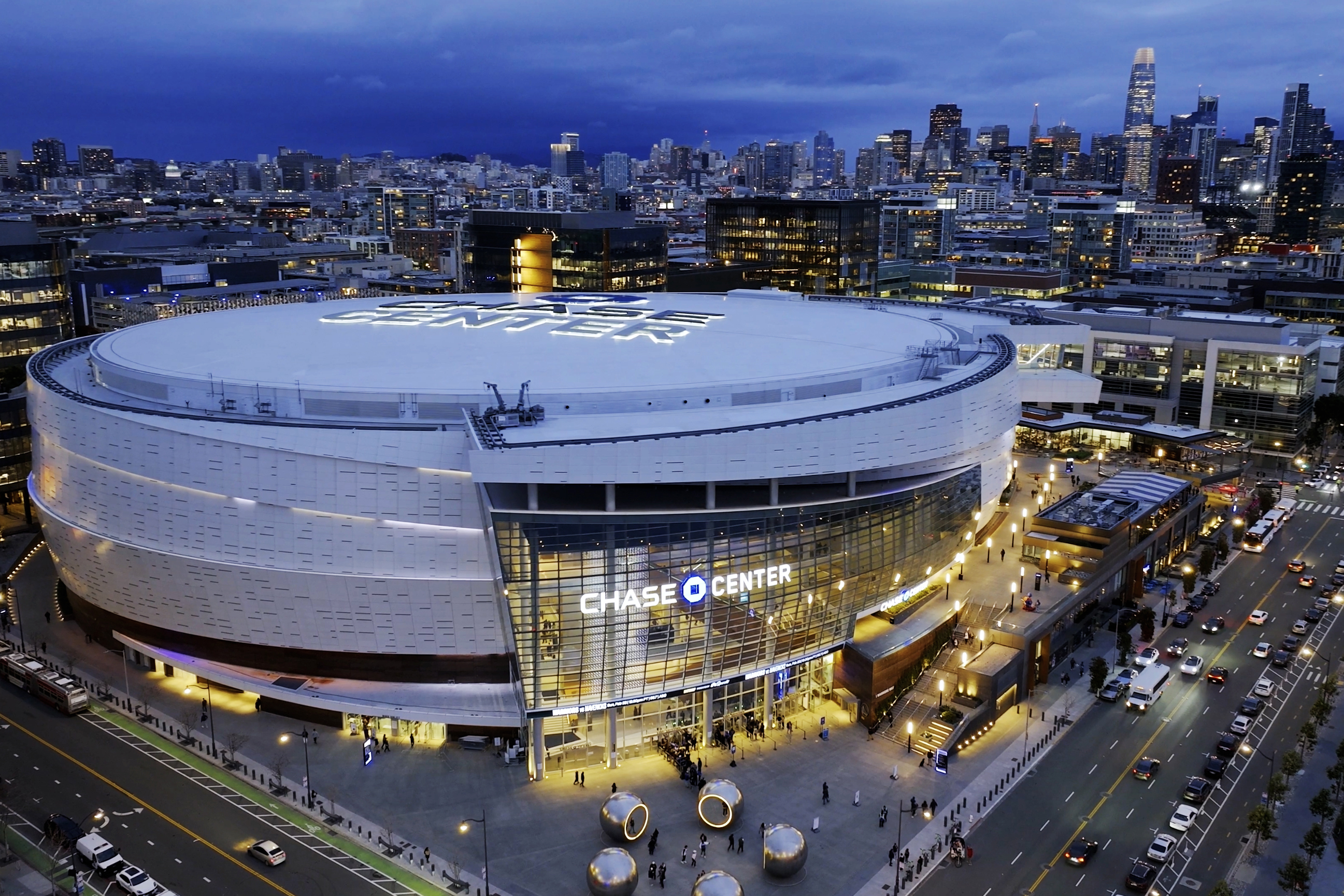 The image shows an illuminated arena with "Chase Center" on the front, surrounded by city buildings, busy streets, and bright lights at dusk.