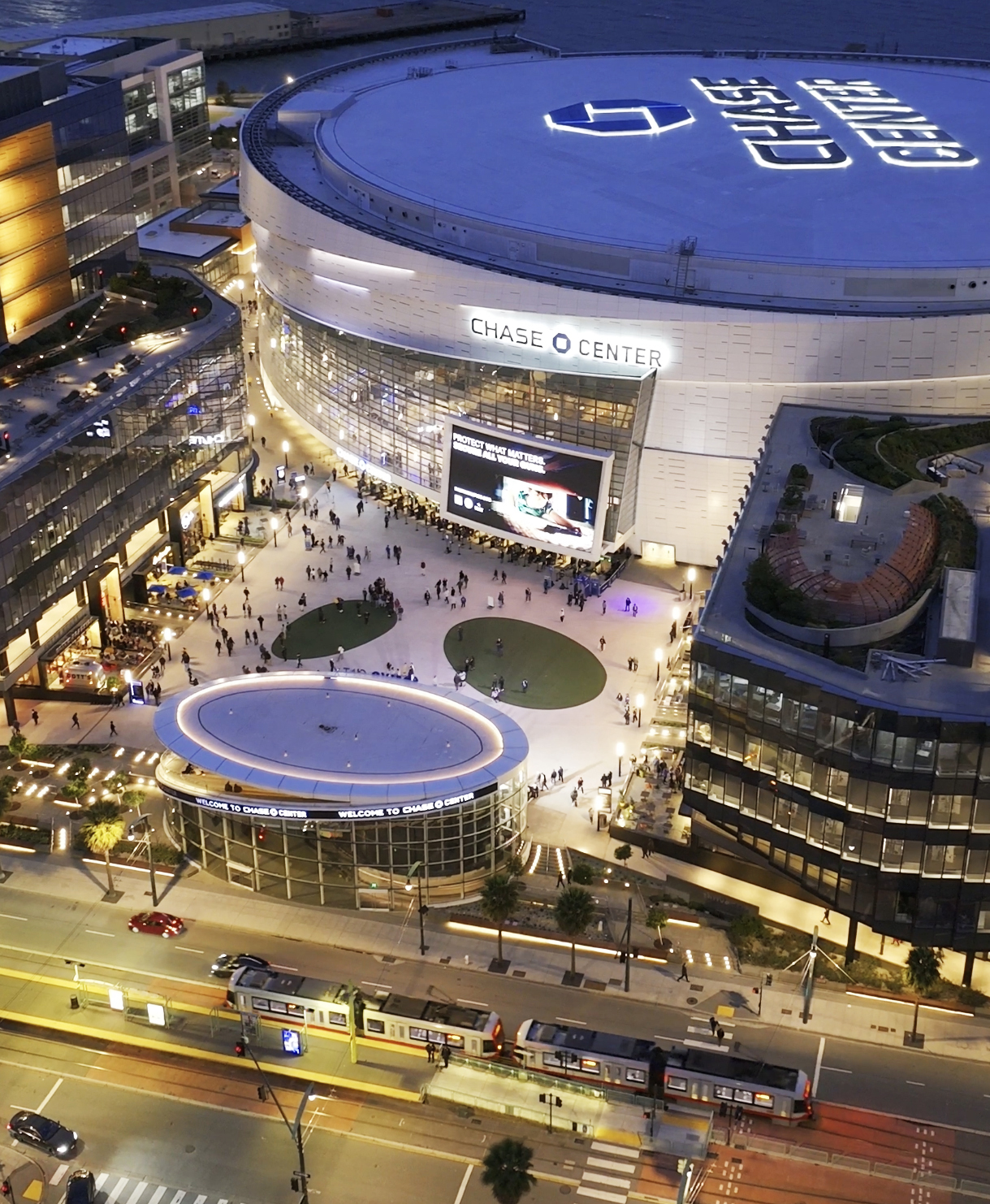 The image shows an aerial view of the Chase Center, a large, circular arena with &quot;CHASE CENTER&quot; written on the roof, surrounded by people and a streetcar.