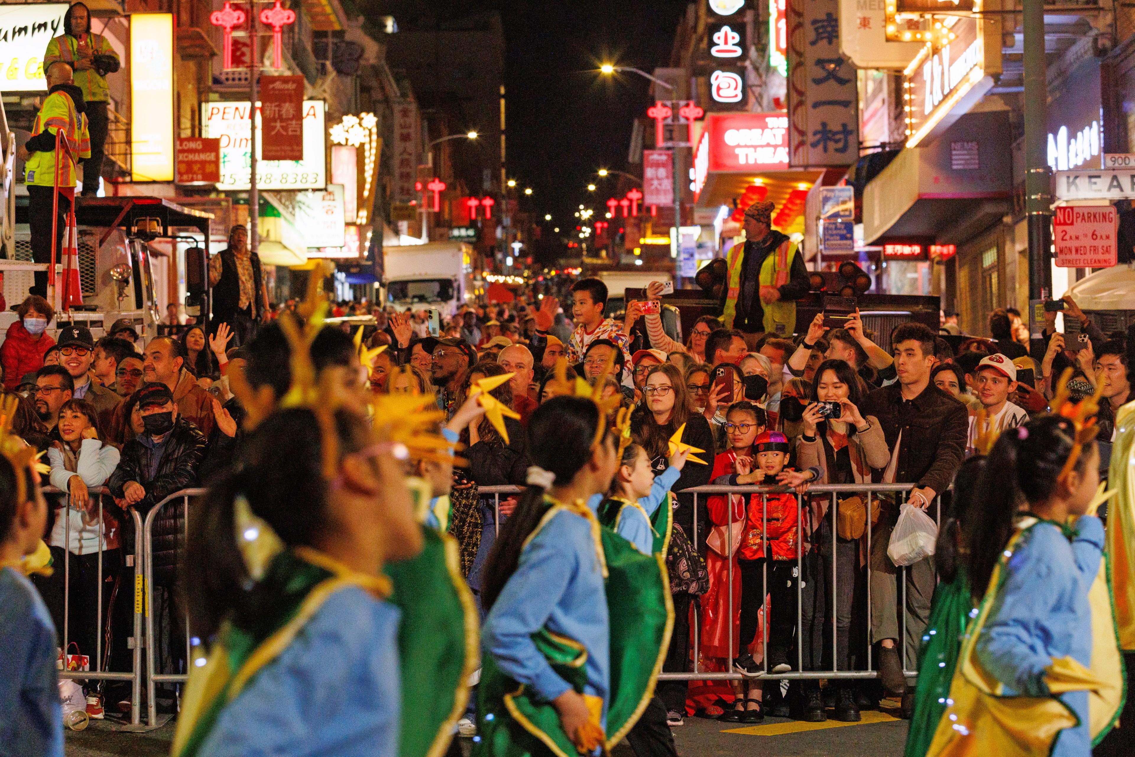 A vibrant street parade at night with dancers wearing colorful costumes. A crowd watches, taking photos, as bright neon signs illuminate the scene.