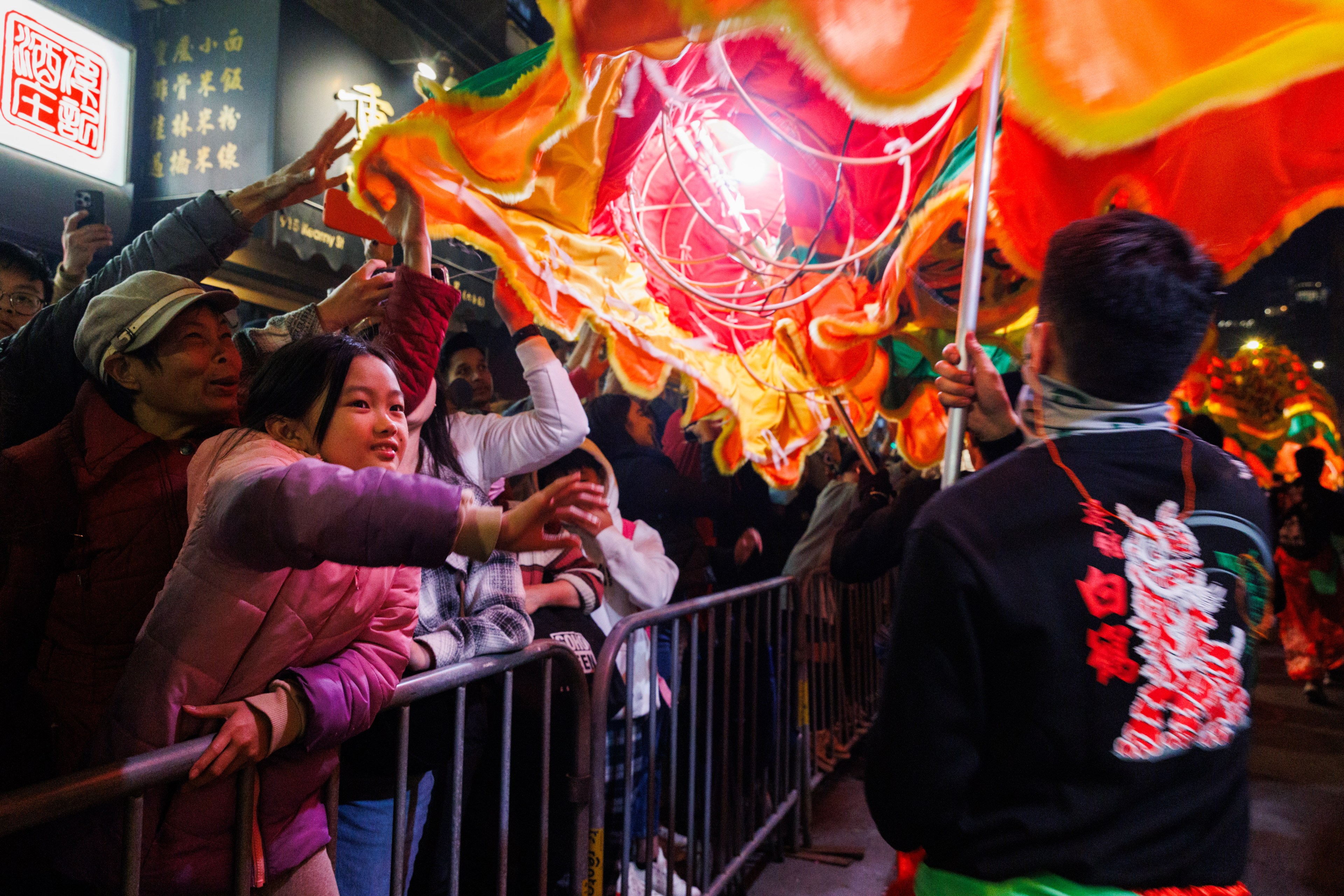 A crowd of excited people reaches out toward a vibrant, multicolored dragon costume in a lively street parade, with bright lights illuminating the scene.