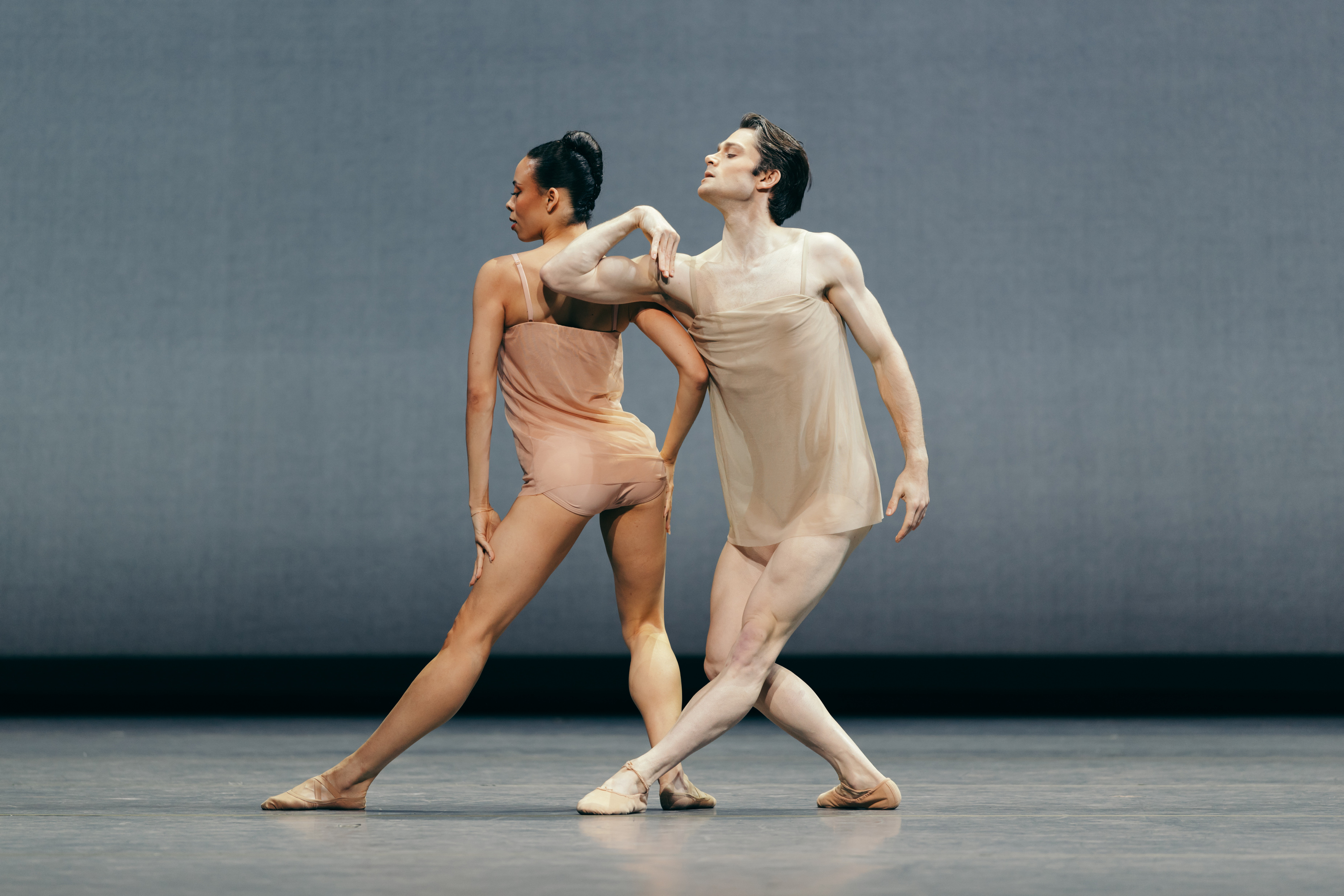 A man and woman gracefully pose in a modern dance performance, wearing sheer, neutral-colored costumes, on a minimalist stage with a plain backdrop.