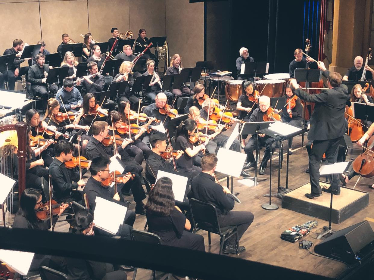 An orchestra is performing on stage, with musicians playing strings, brass, woodwinds, and percussion. The conductor stands on a podium guiding them.