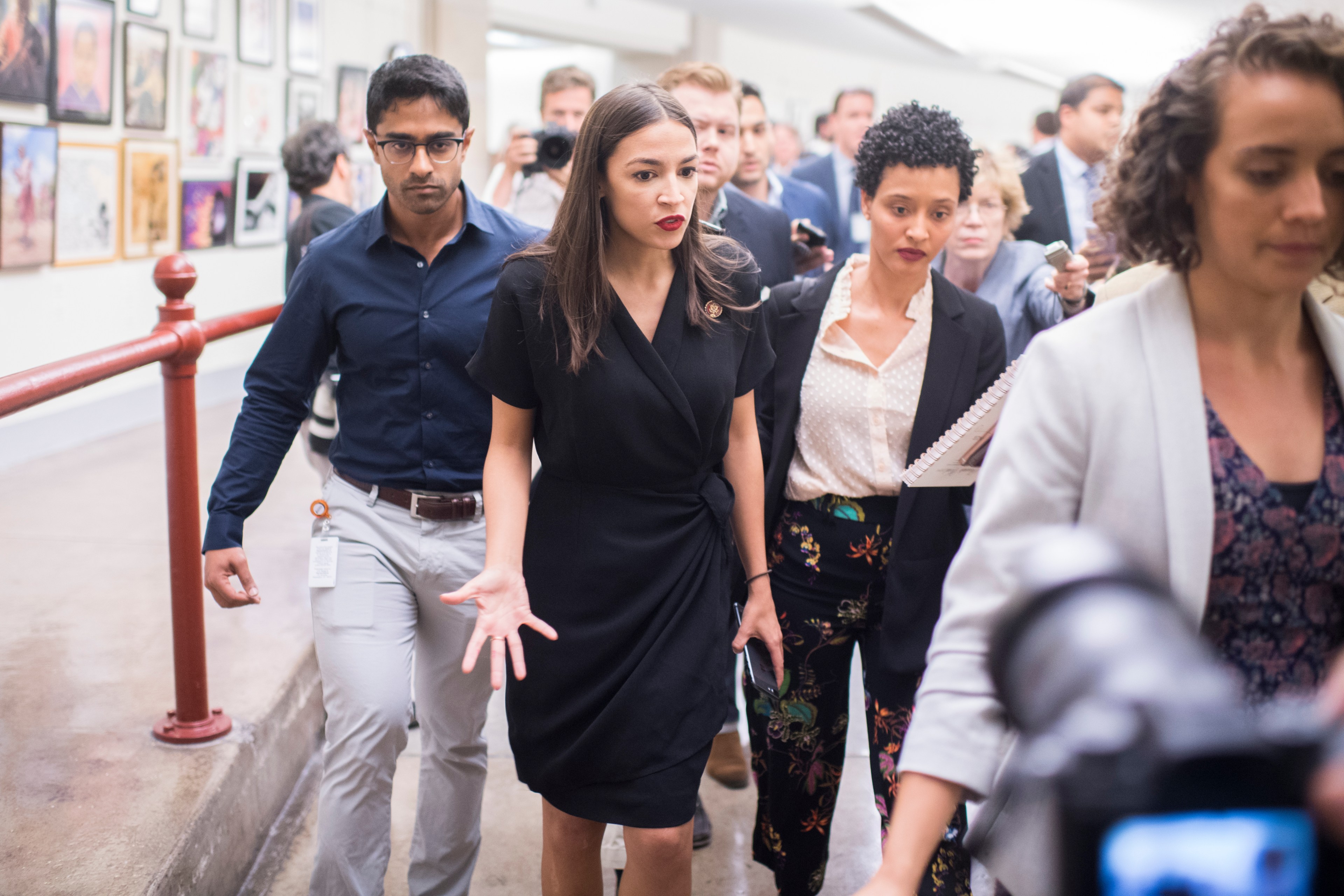 A group of people walk together in a hallway. The woman in front wears a black dress, flanked by individuals in business attire. Other people and cameras are visible.