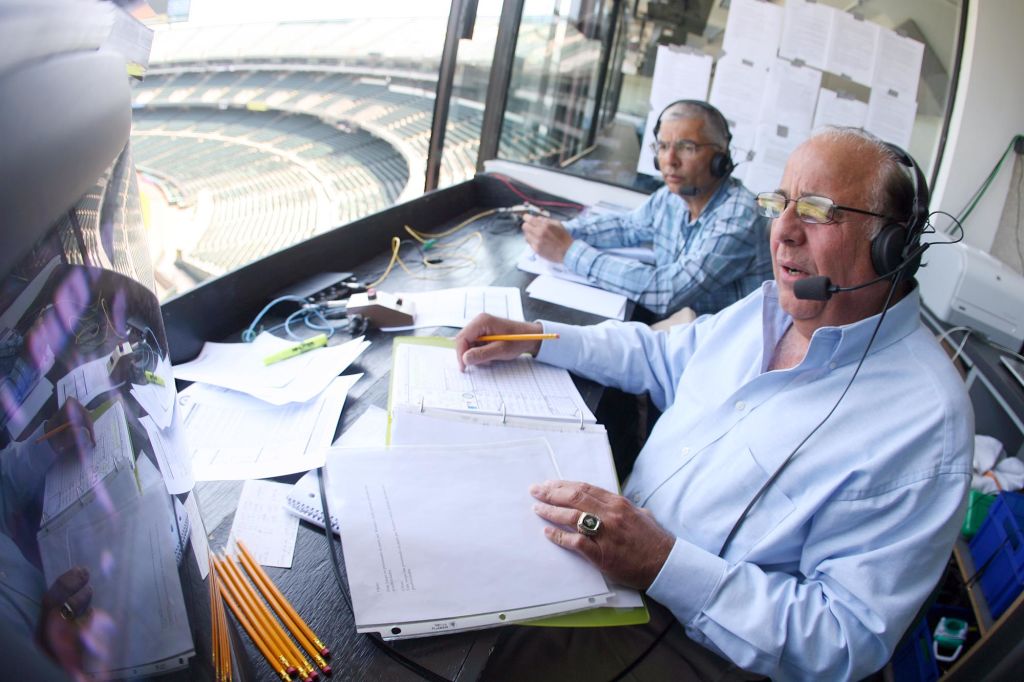 Two broadcasters in headphones sit in a booth overlooking an empty stadium, with papers and pencils on the desk. One is speaking into a microphone.