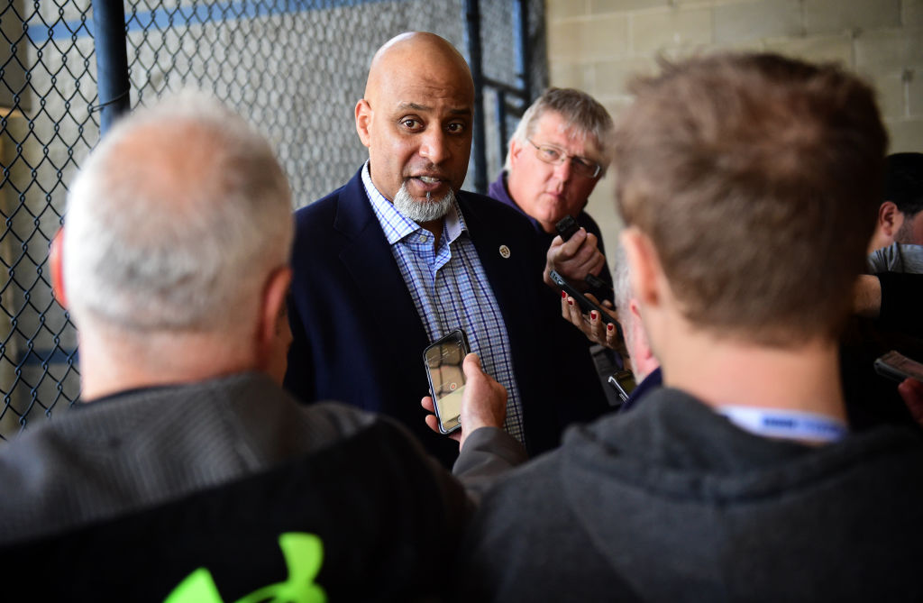 A man in a suit speaks to several reporters holding microphones. They are standing near a chain-link fence, and one reporter wears a jacket with a green emblem.