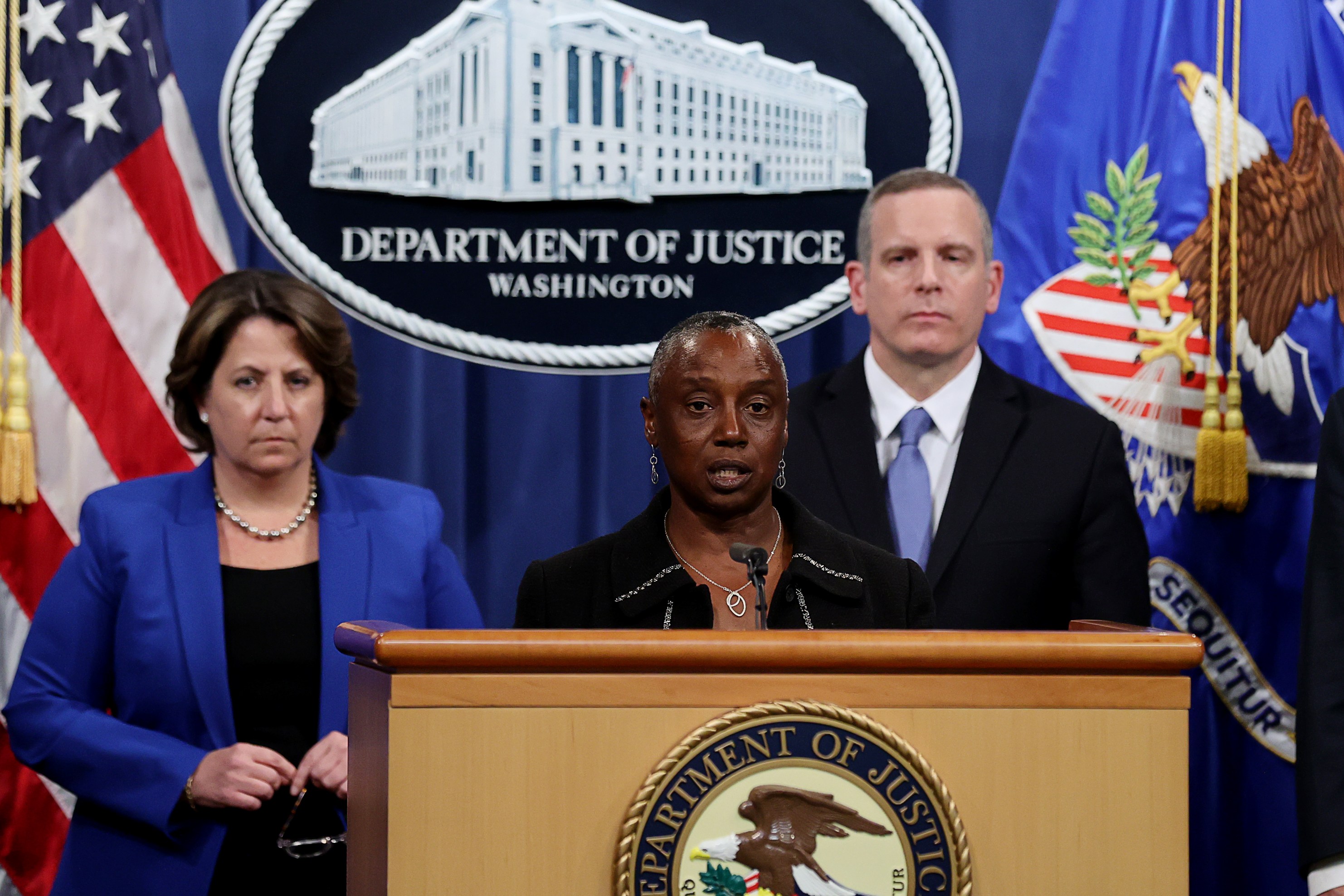 Three officials stand at a podium with the Department of Justice seal. The woman in the center speaks, flanked by a woman in blue and a man in a suit.