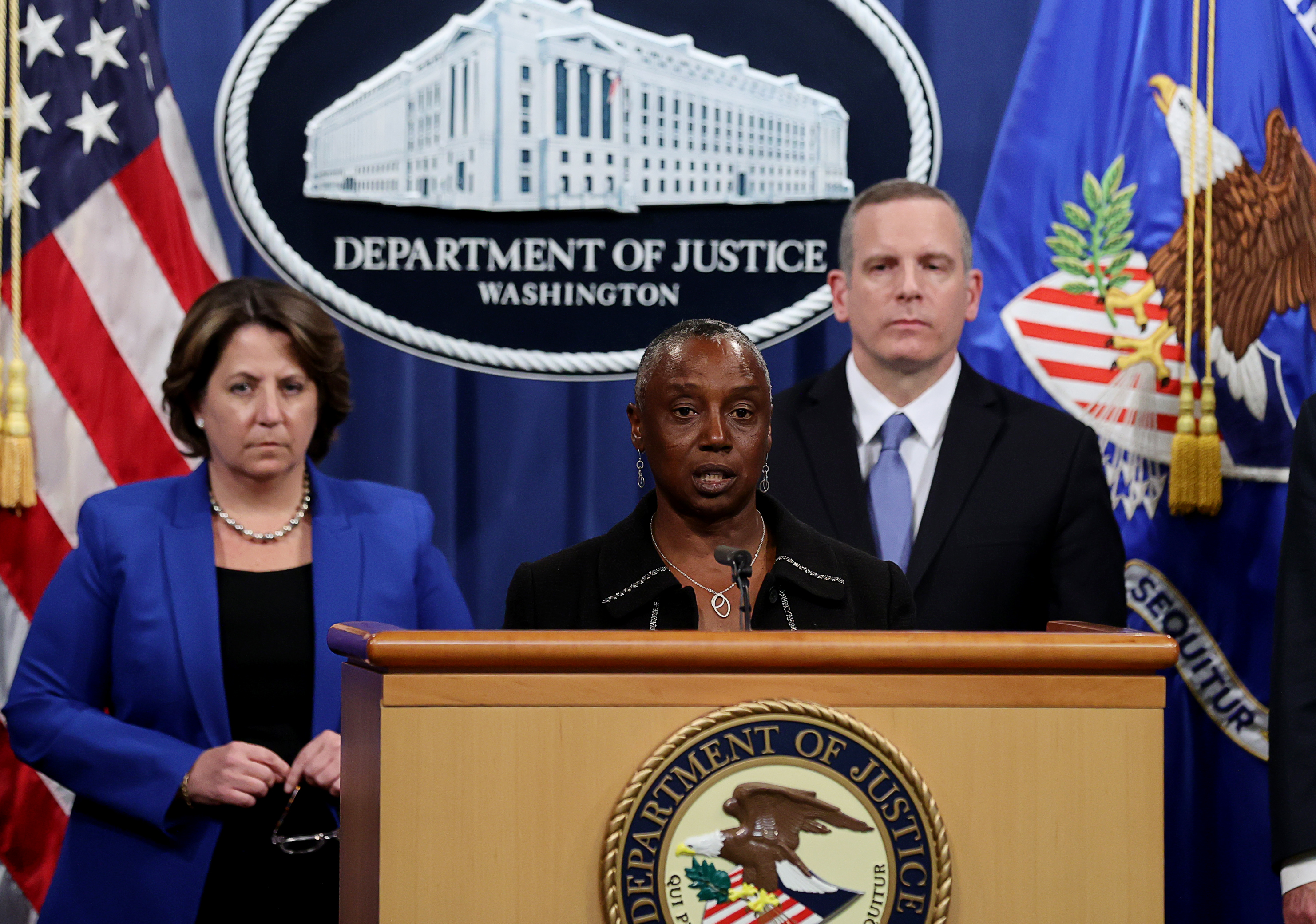 Three officials stand at a podium with the Department of Justice seal. The woman in the center speaks, flanked by a woman in blue and a man in a suit.