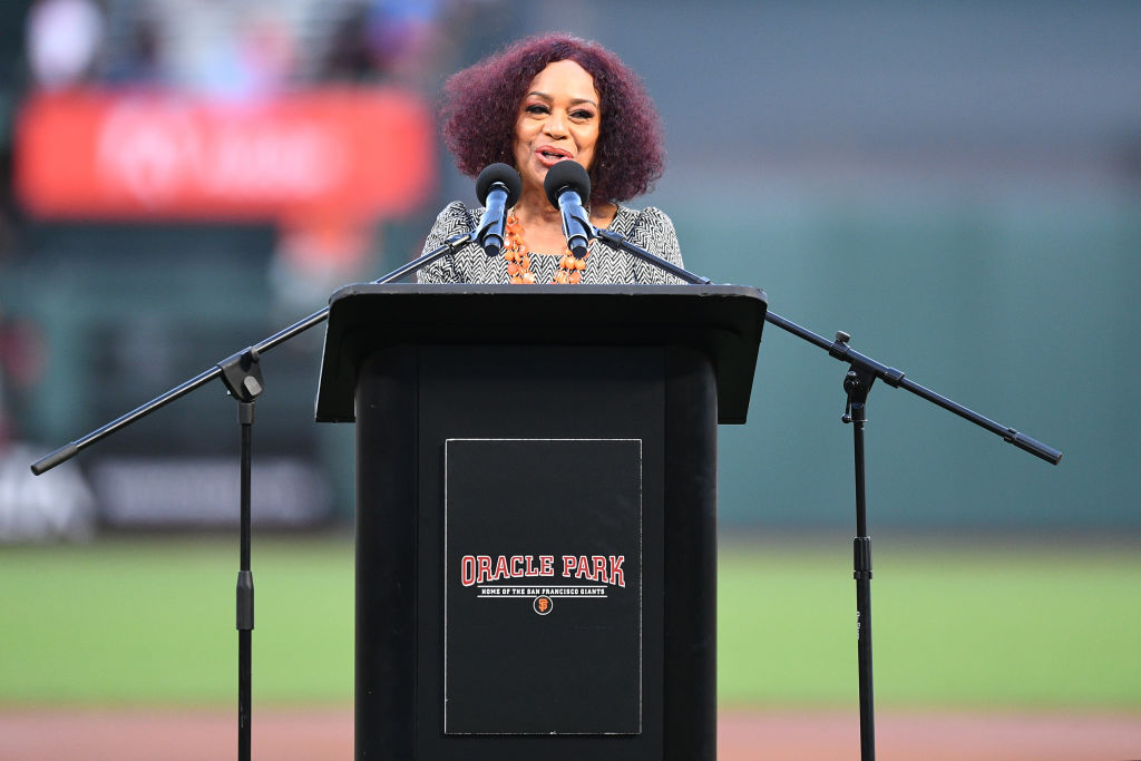 A person stands at a podium labeled "Oracle Park" with two microphones. The background is a blurred view of a sports field.