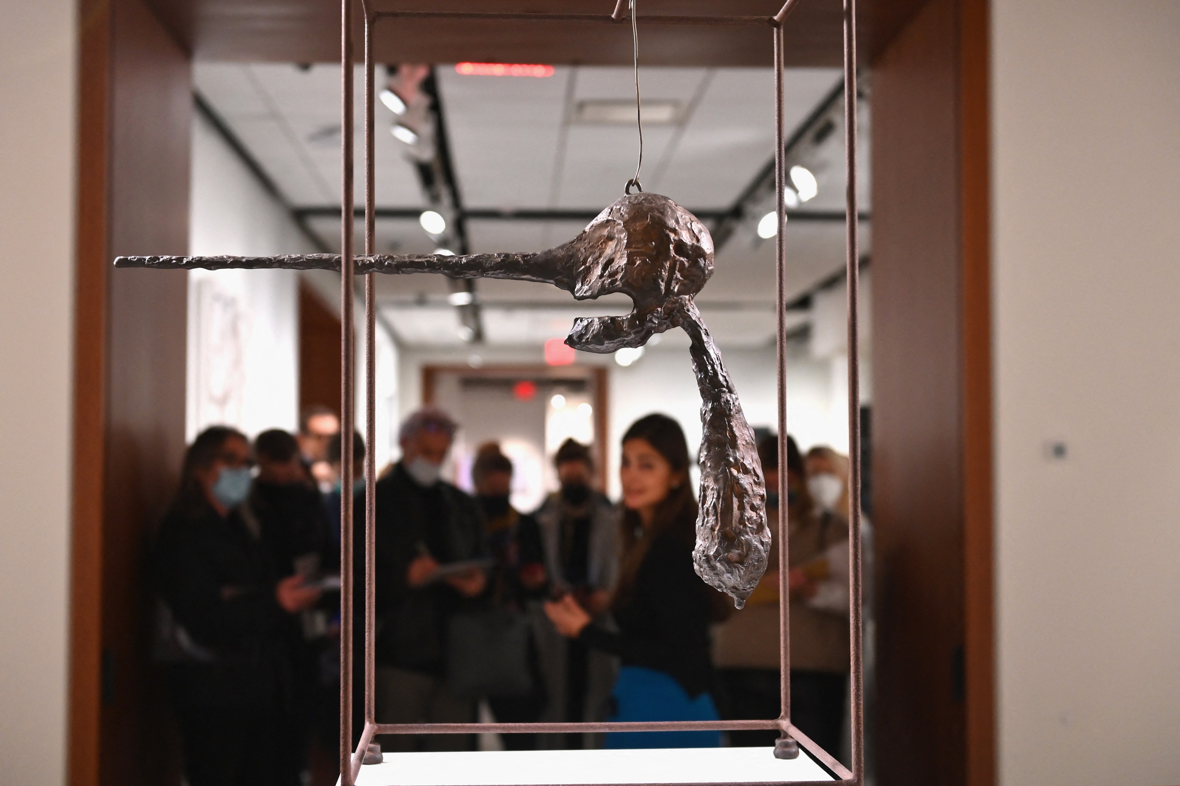 A metallic abstract sculpture is suspended in the foreground, with a group of people in the background, some wearing masks, observing attentively.