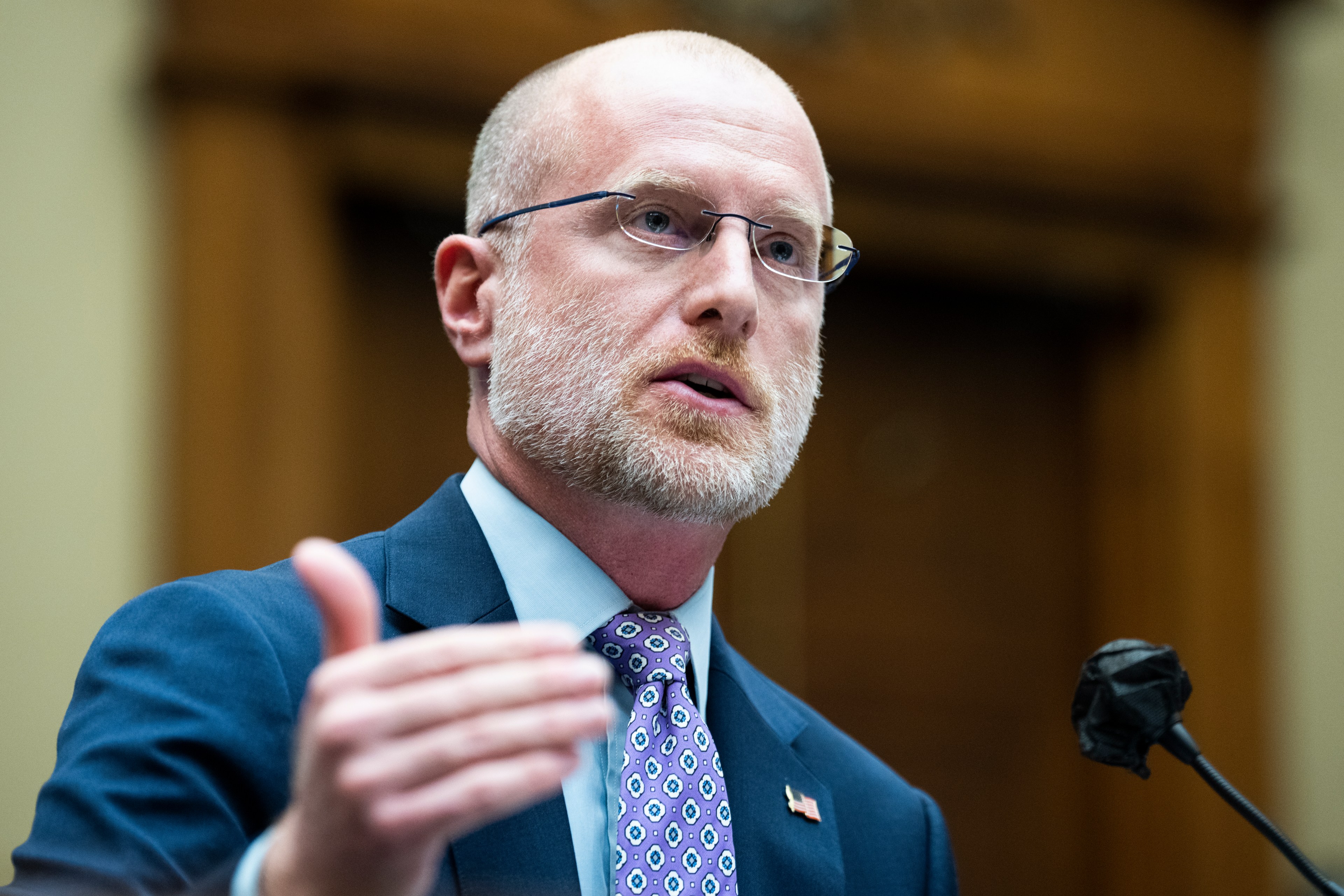 A man with glasses and a beard speaks, gesturing with his hand. He's wearing a blue suit, light blue shirt, and patterned tie. The background is blurred.